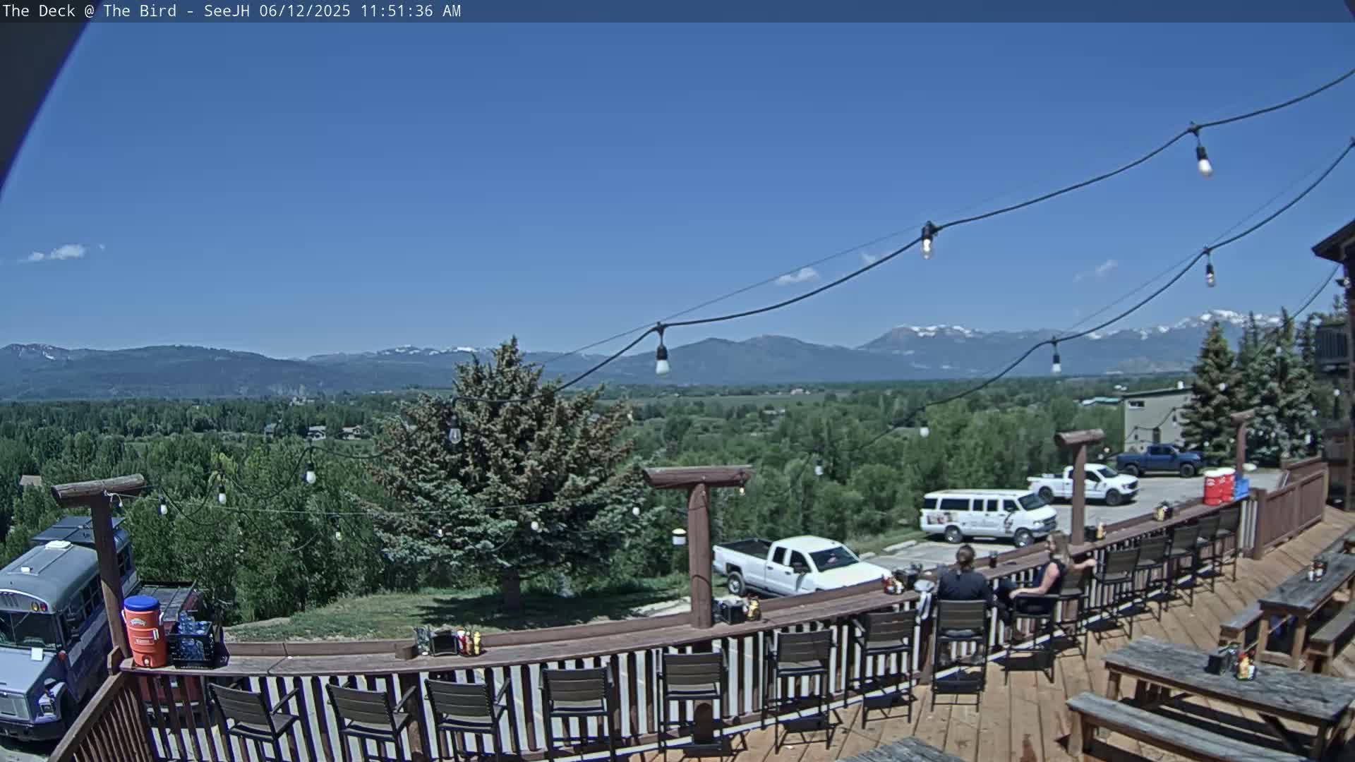 An outdoor deck with seating overlooks a valley, mountains, and parked vehicles under a clear blue sky.