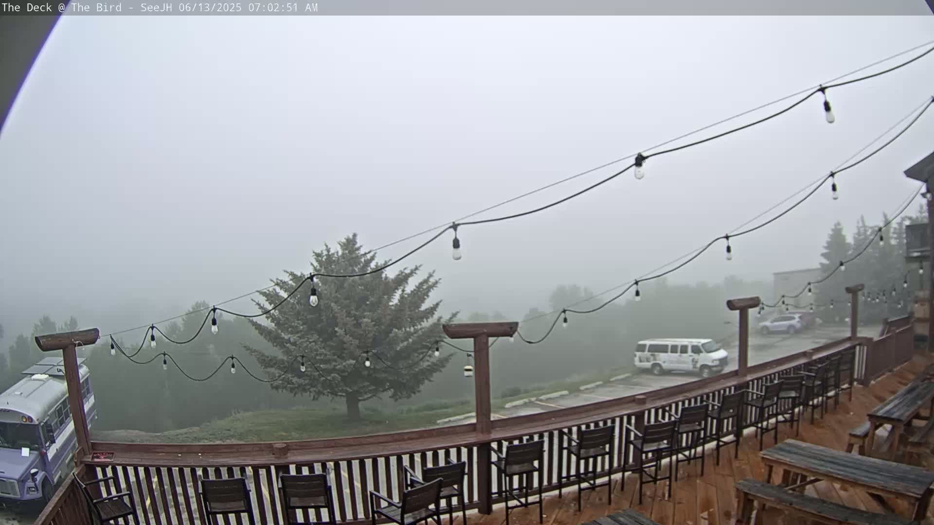 A foggy outdoor deck with string lights overlooks a parking lot and a bus, with several empty chairs and picnic tables on the deck.