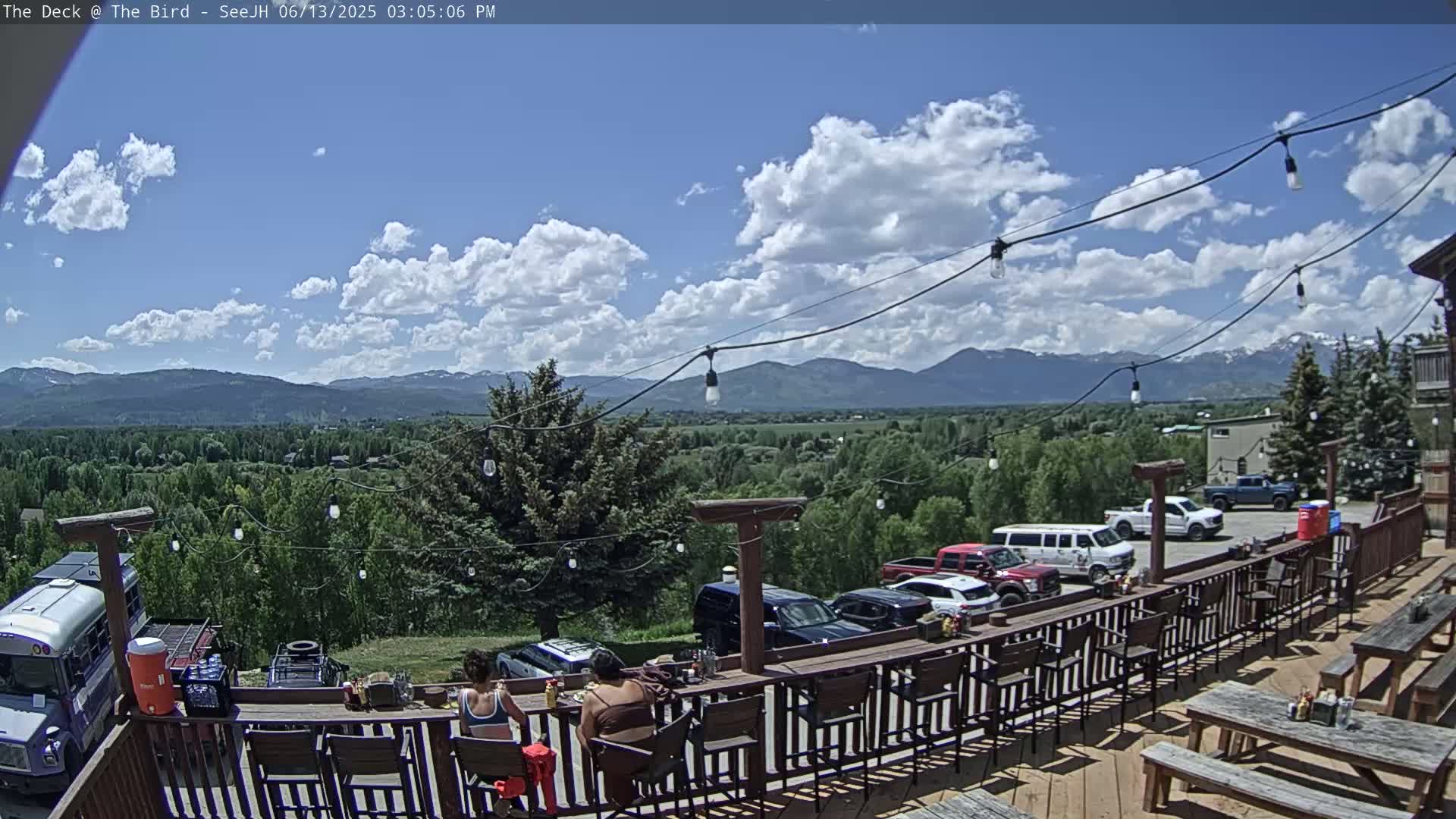 An outdoor deck with a bar and seating overlooks a valley, mountains, and a parking lot under a partly cloudy sky.