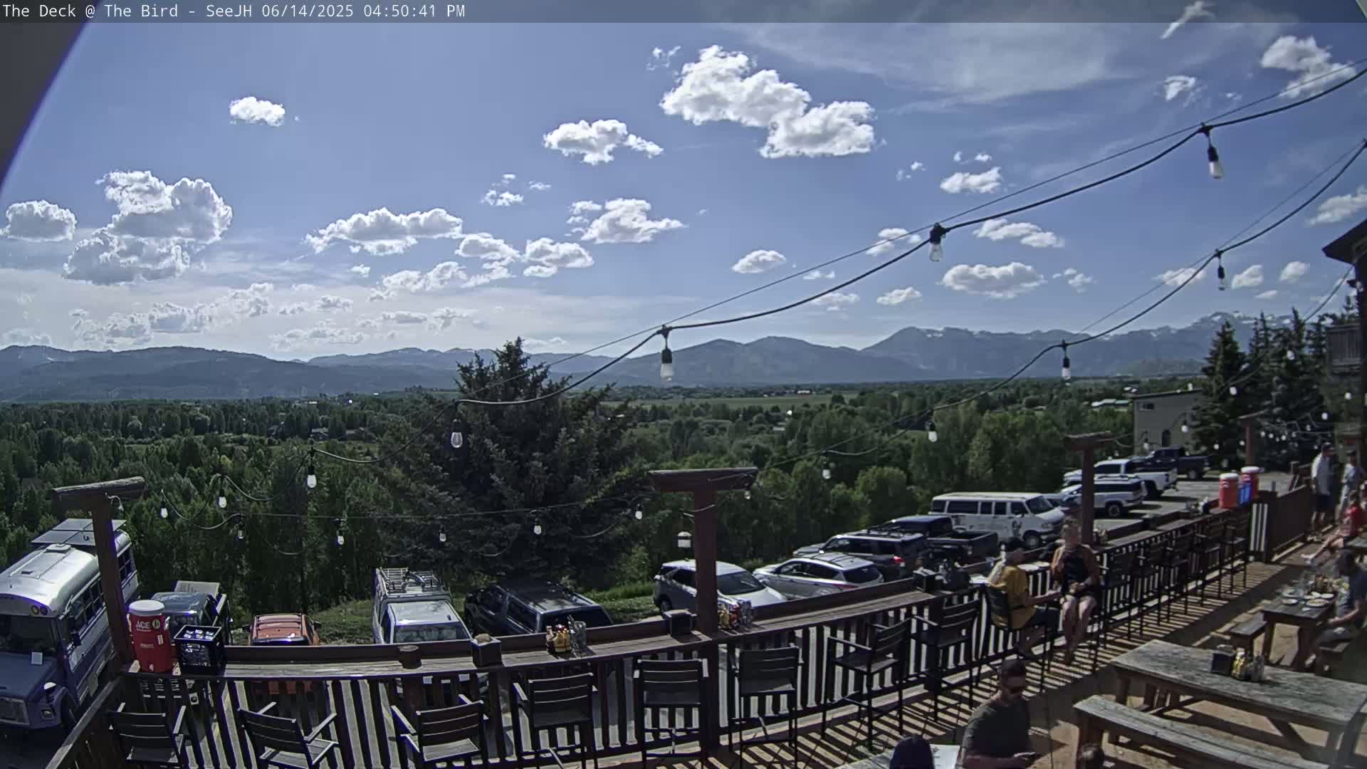 An outdoor deck with a bar and seating overlooks a valley and distant mountains under a partly cloudy sky.