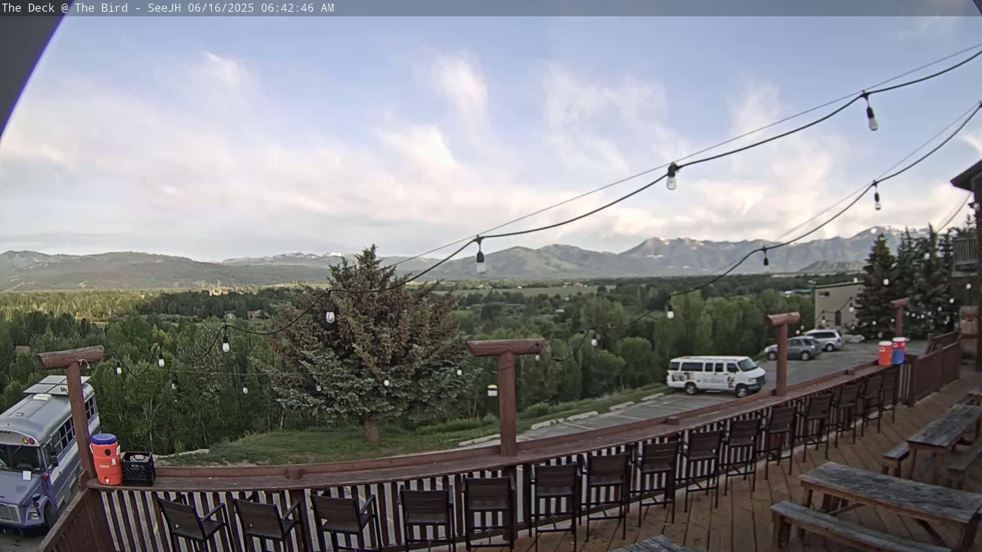A deck with chairs and picnic tables overlooks a valley with mountains in the distance under a partly cloudy sky.