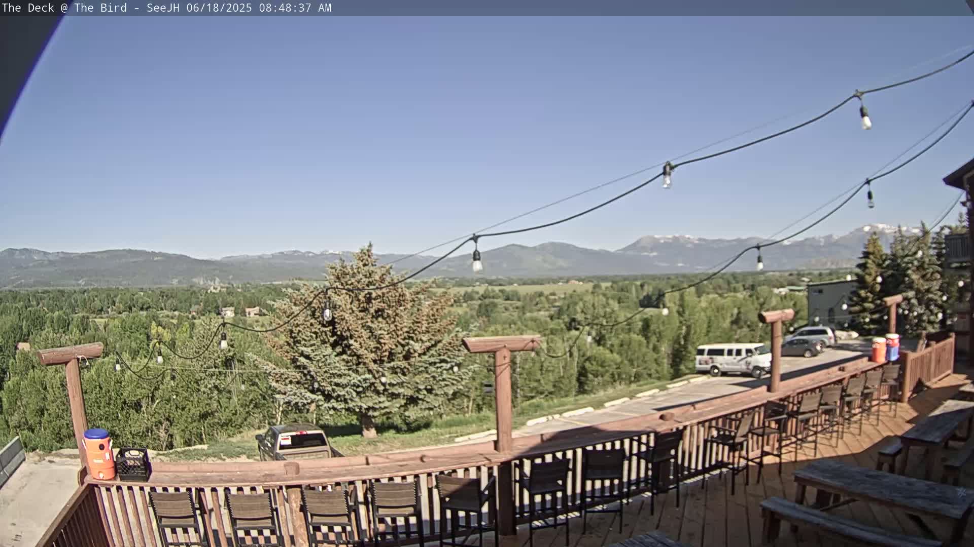 A deck with wooden railings and chairs overlooks a valley of green trees and distant mountains under a clear blue sky.