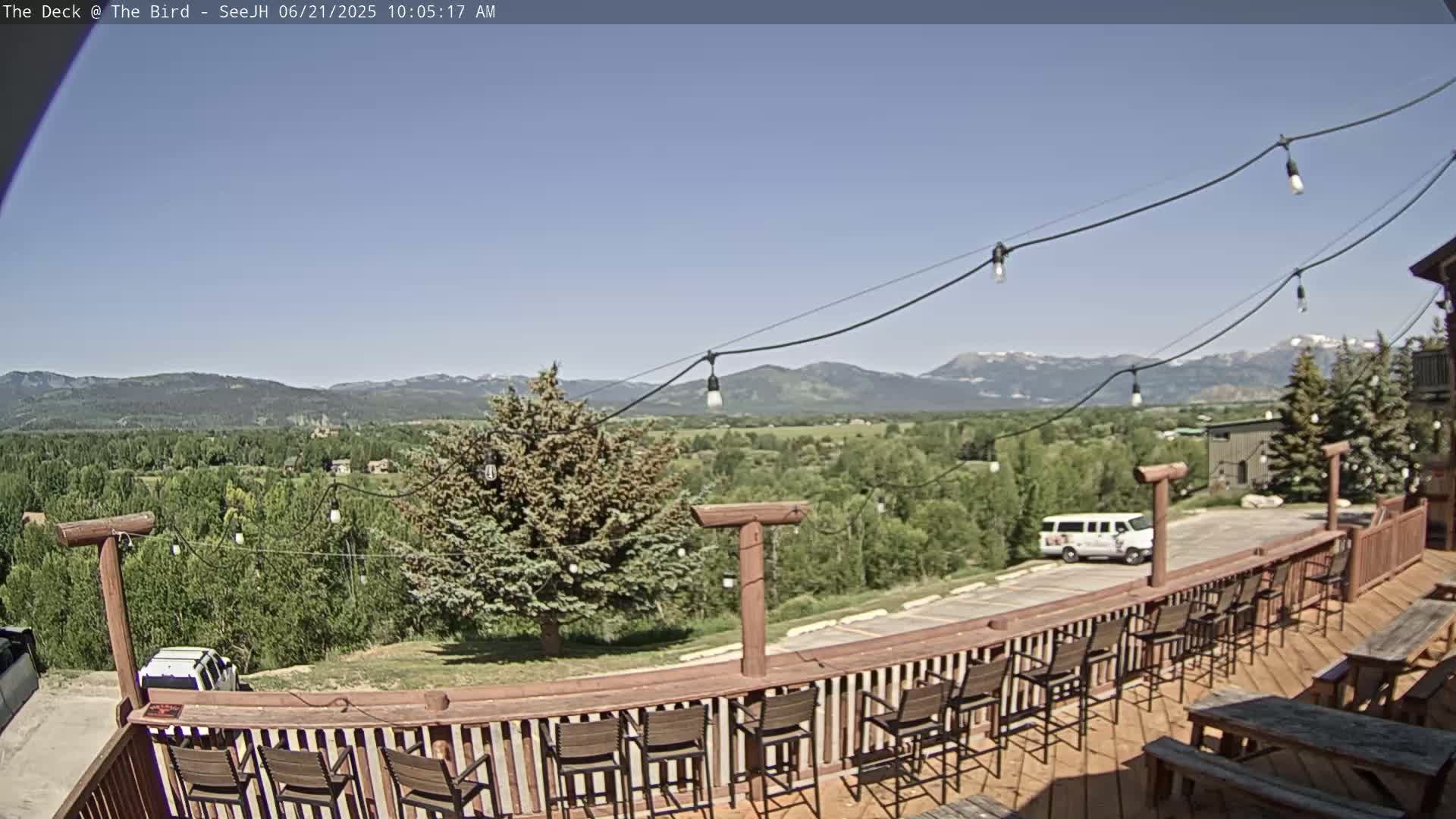 A wooden deck with chairs and tables overlooks a valley of green trees and distant mountains under a clear blue sky.