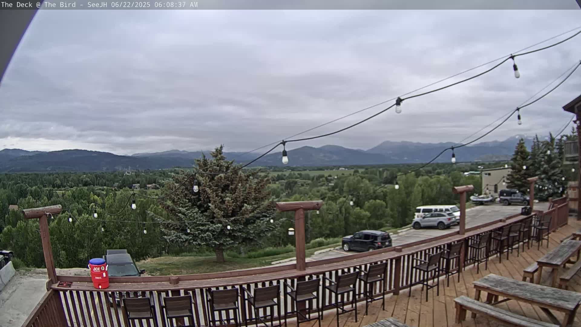 A wooden deck with bar stools and picnic tables overlooks a valley and distant mountains under a cloudy sky.