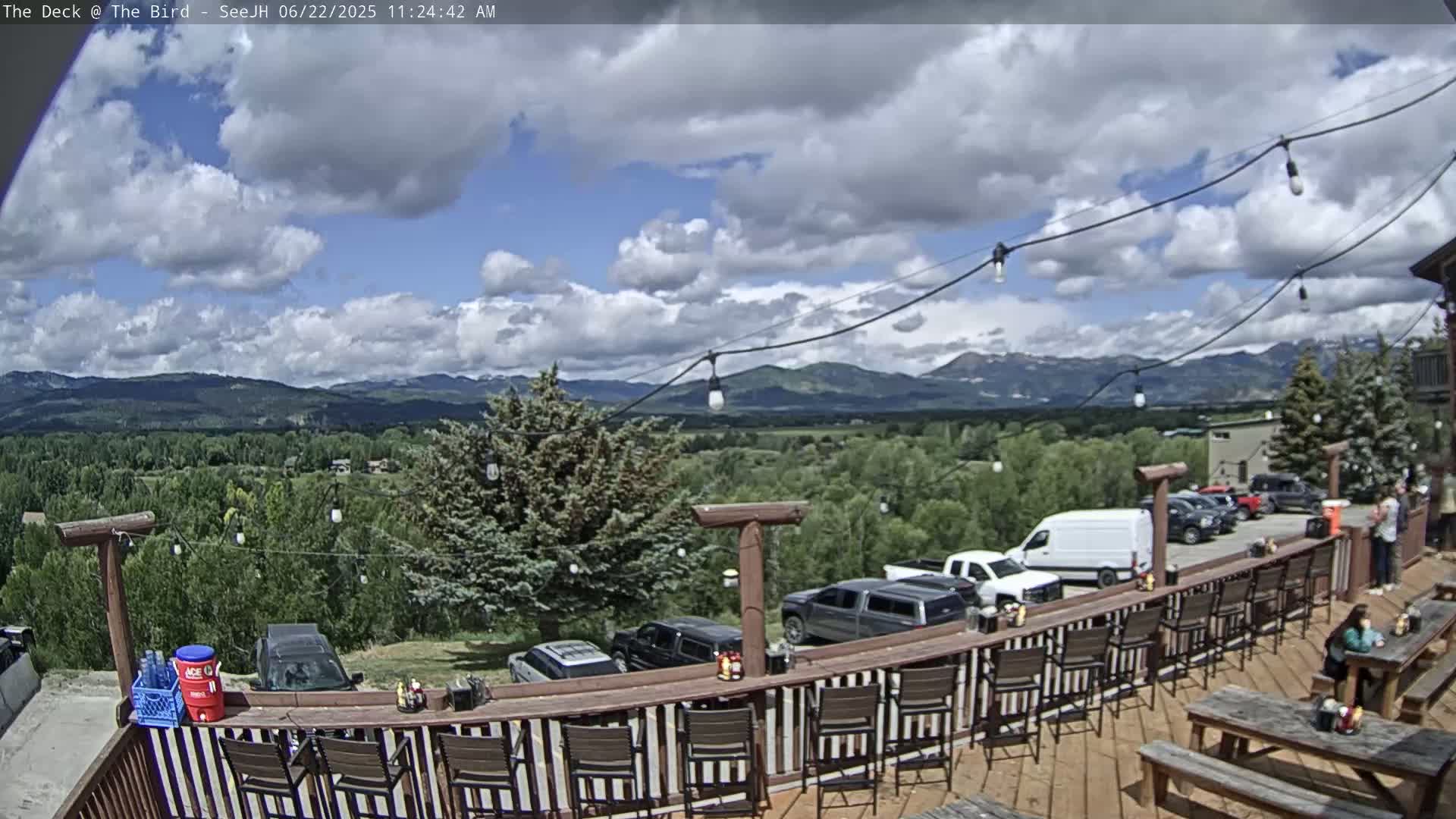 An outdoor deck with a bar and picnic tables overlooks a valley and mountains under a partly cloudy sky.