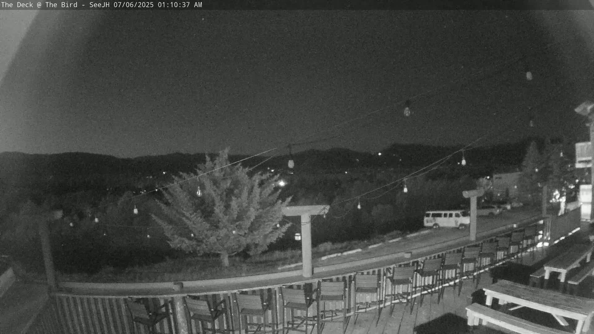A nighttime outdoor scene shows a deck with empty chairs and picnic tables overlooking a tree and distant hills under a clear sky.