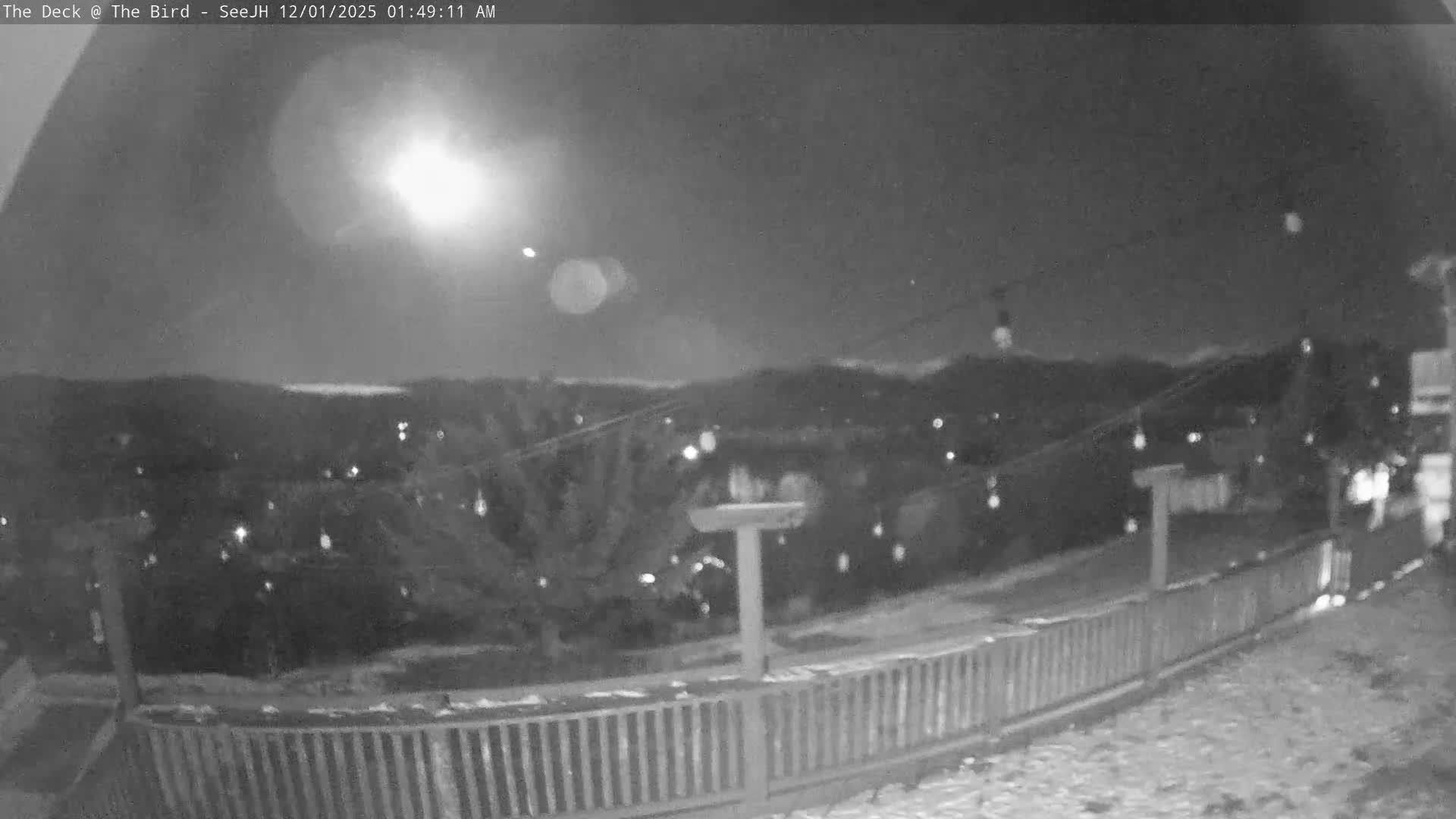 A monochrome night scene depicts a very bright light source in the sky above a snowy landscape with scattered distant lights, viewed over a wooden deck railing, under clear and cold conditions.