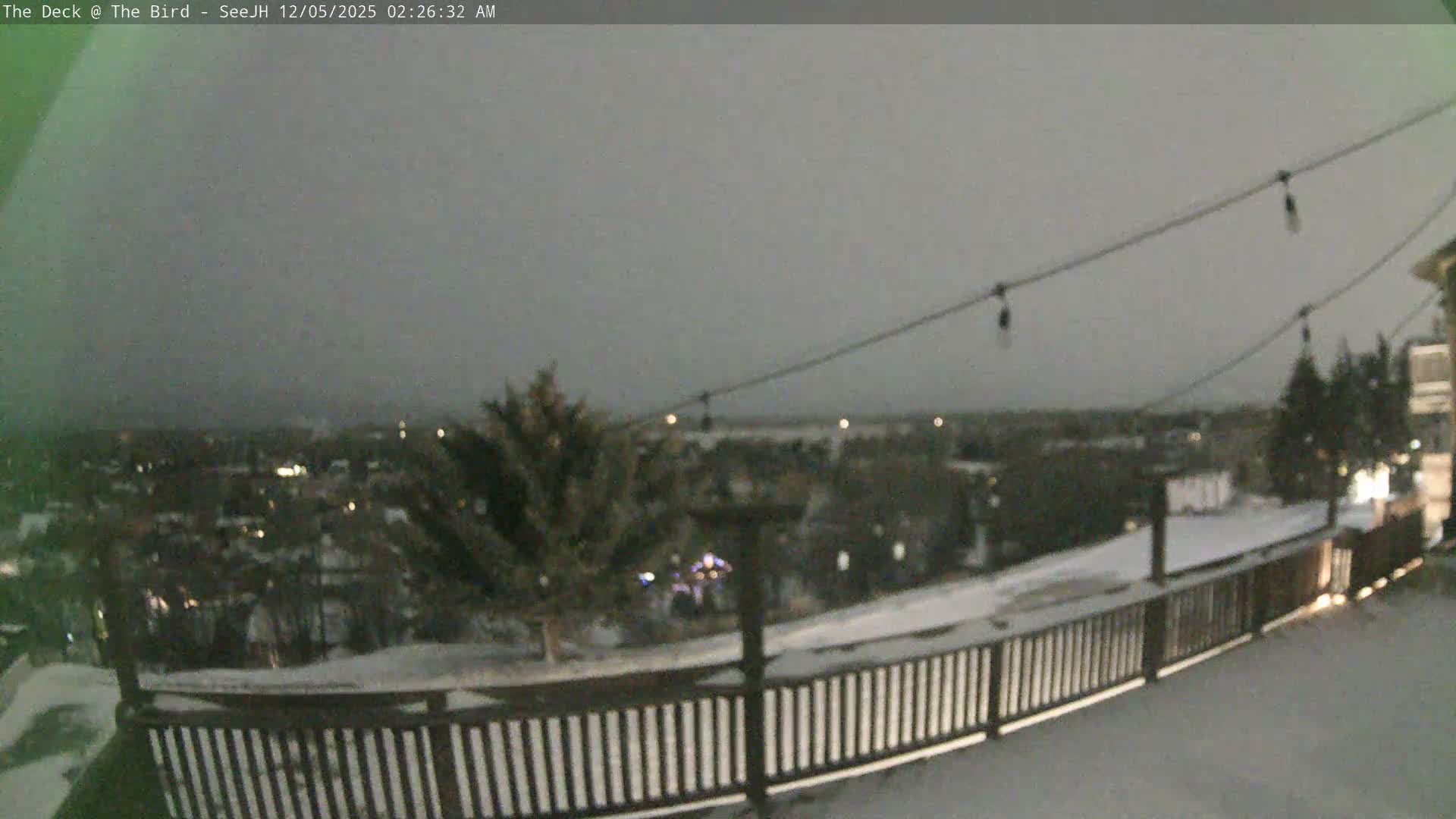 A snow-covered outdoor deck with string lights extends into the foreground, offering a view of a sprawling, dark landscape dotted with distant lights under an overcast winter sky.