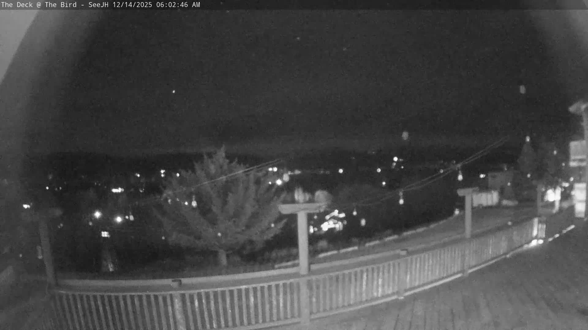 A snow-covered outdoor deck with string lights extends into the foreground, offering a view of a sprawling, dark landscape dotted with distant lights under an overcast winter sky.