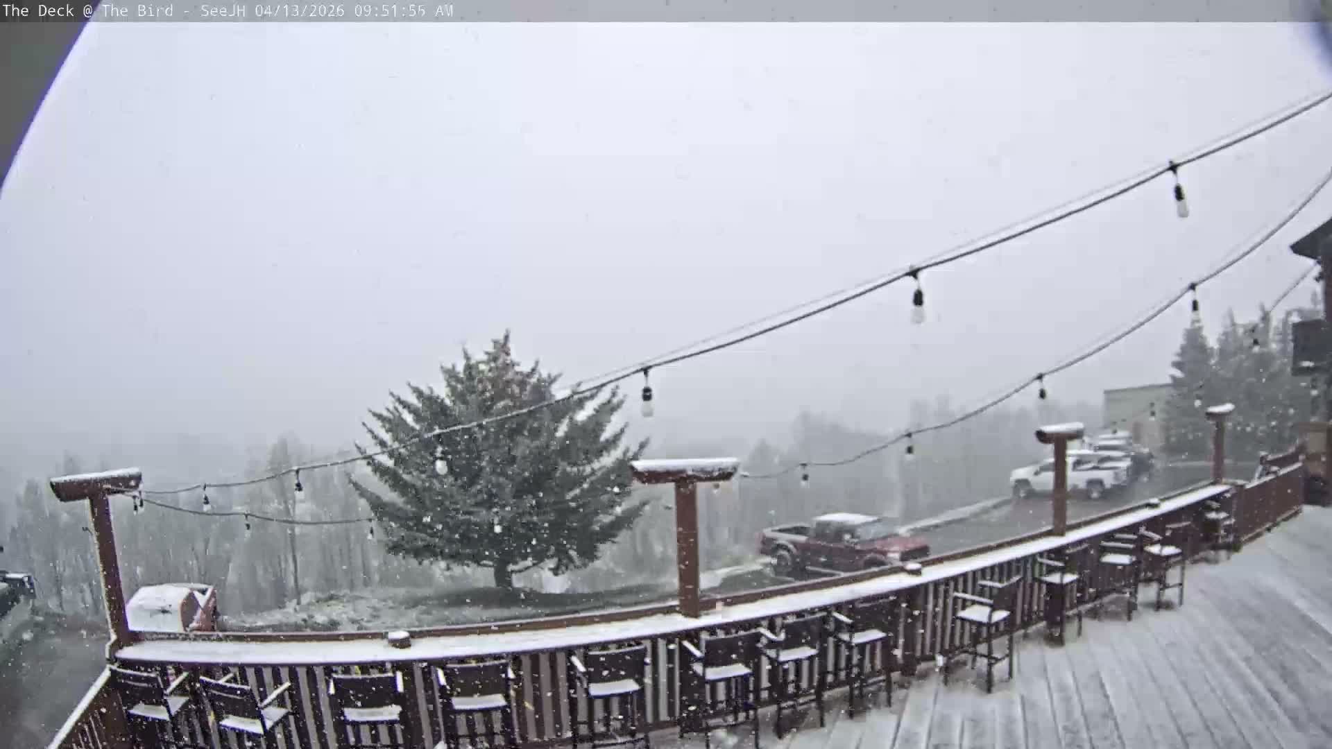 A snow-covered outdoor deck with string lights extends into the foreground, offering a view of a sprawling, dark landscape dotted with distant lights under an overcast winter sky.