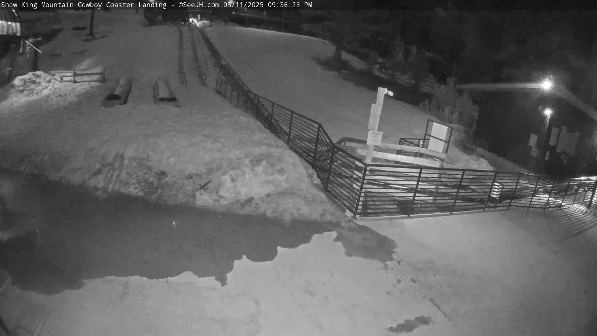 A snow-covered area at night, featuring a metal fence, a small building, and a dark puddle of water.