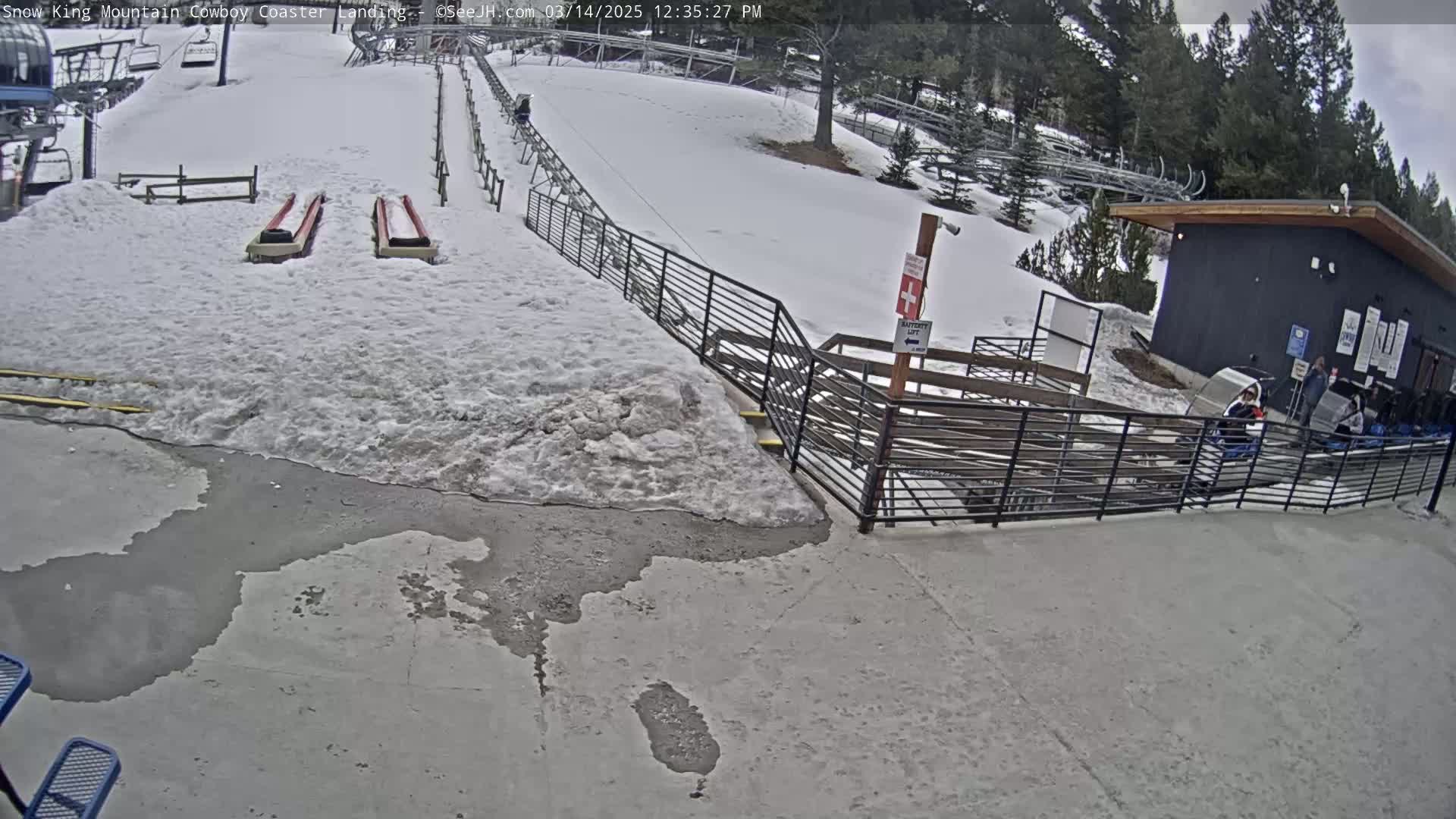 A snow-covered area with two empty sledding runs, a small building, and metal railings, with some melting snow and puddles on the concrete ground.
