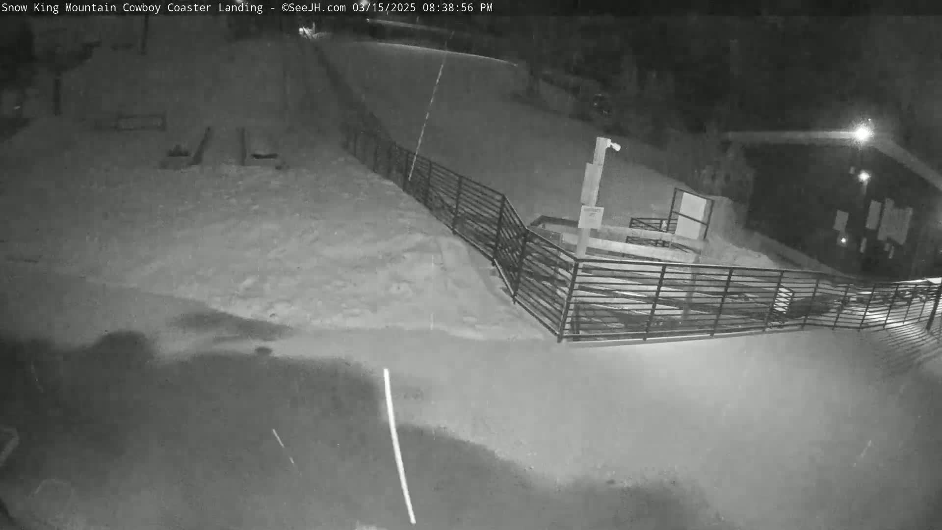 A snow-covered area at night, illuminated by artificial light, features a metal fence and a building in the background, with light snow falling.