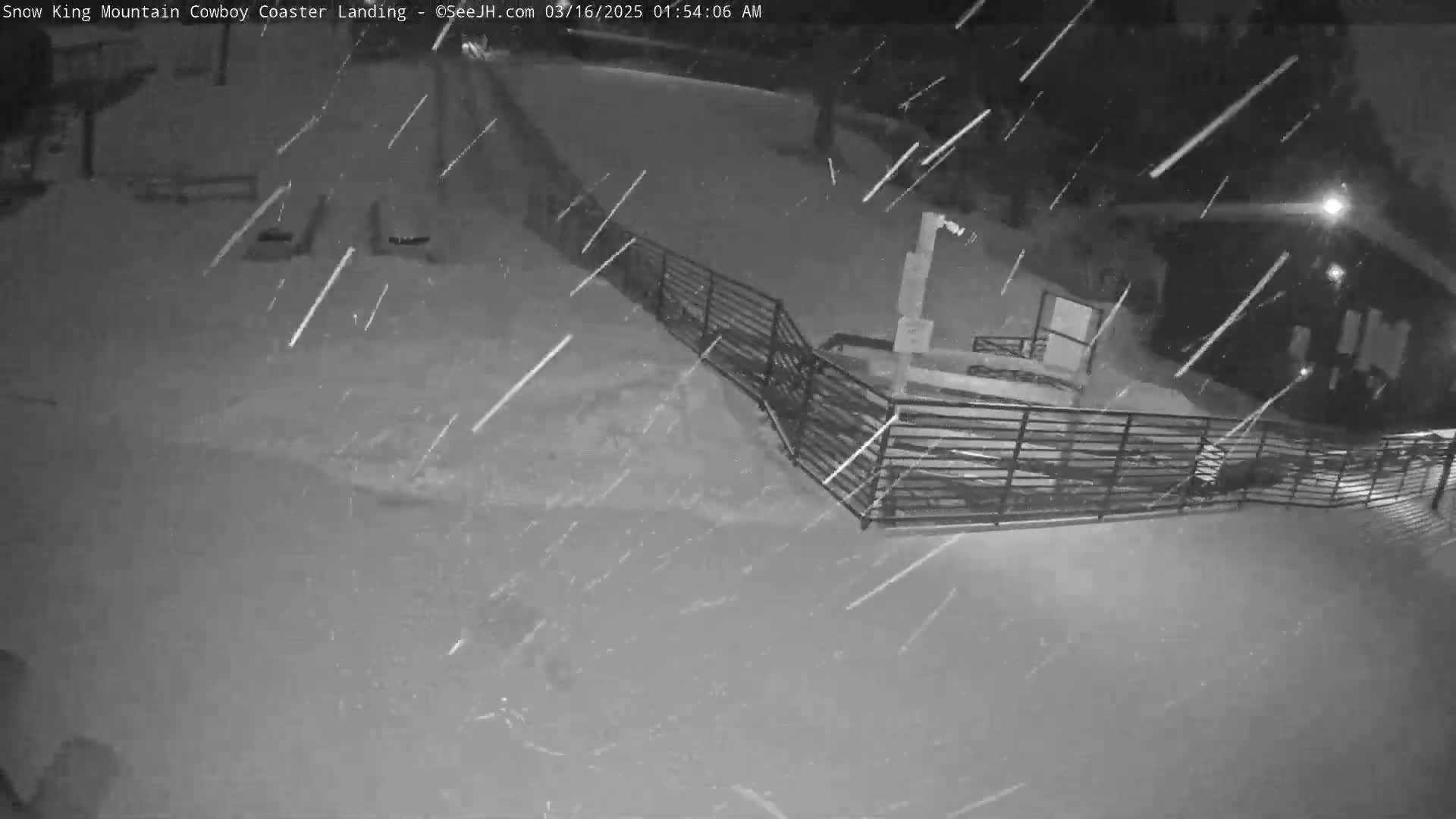A snow-covered area with a metal fence and building is seen under heavy snowfall at night.