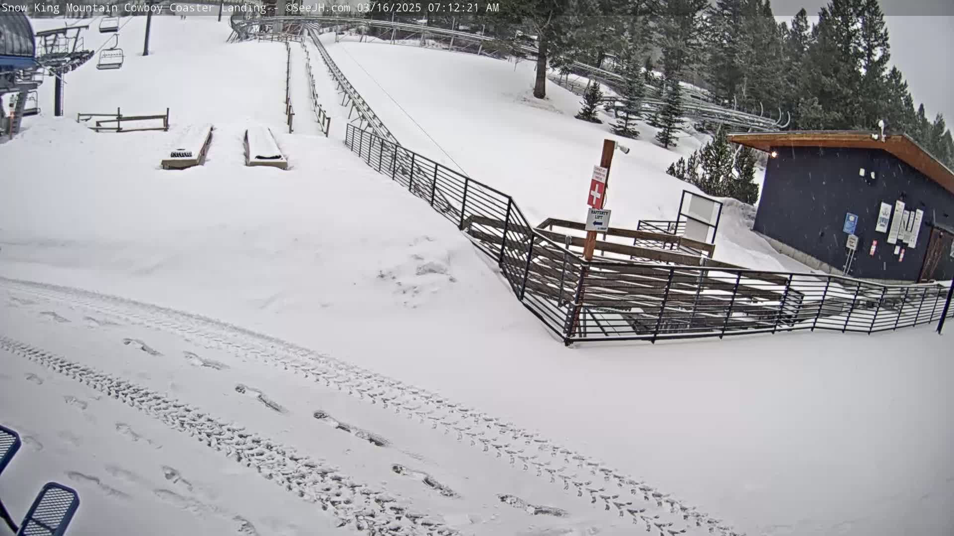 A snow-covered area with a coaster landing, a small building, and footprints in the snow during a snowfall.
