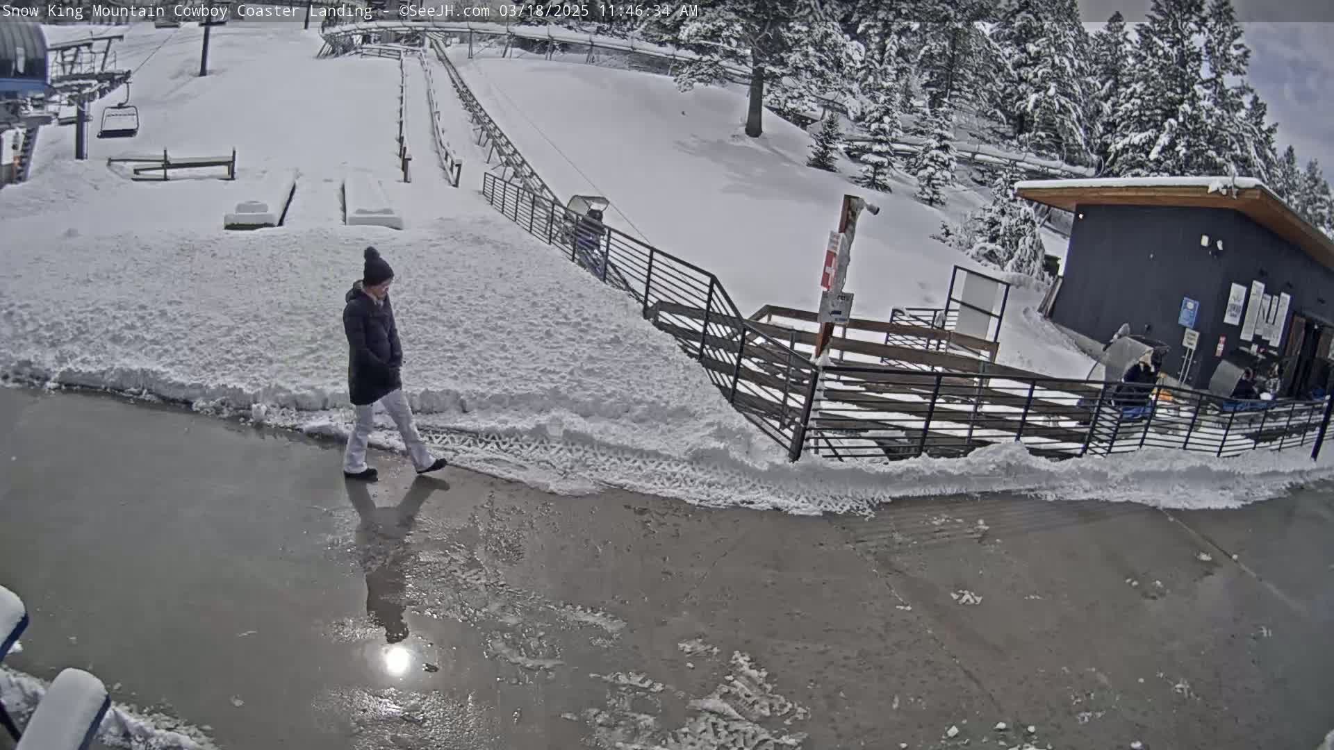 A person walks on a snow-covered paved area near a snow-covered mountainside, a building, and a metal fence, in snowy conditions.