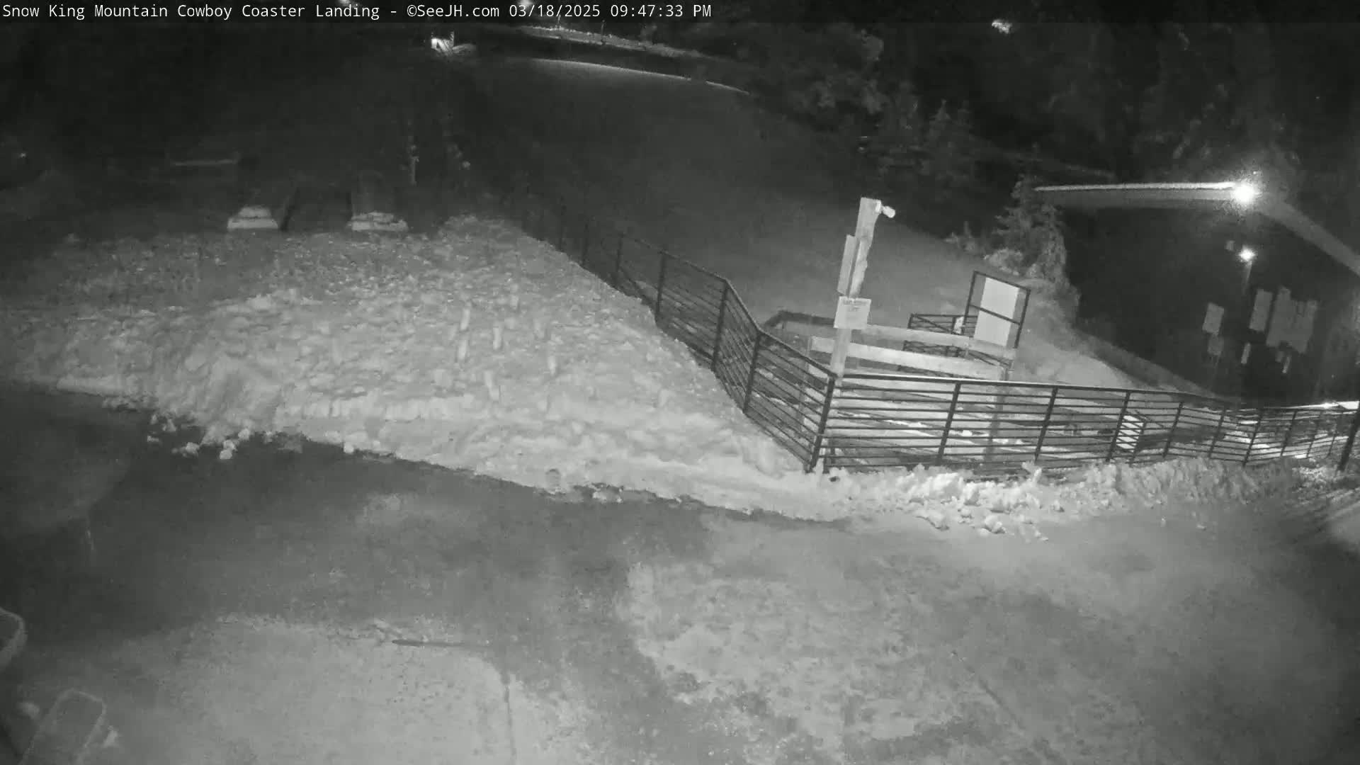 A nighttime, snow-covered area with a metal fence and a small building is visible under artificial light.