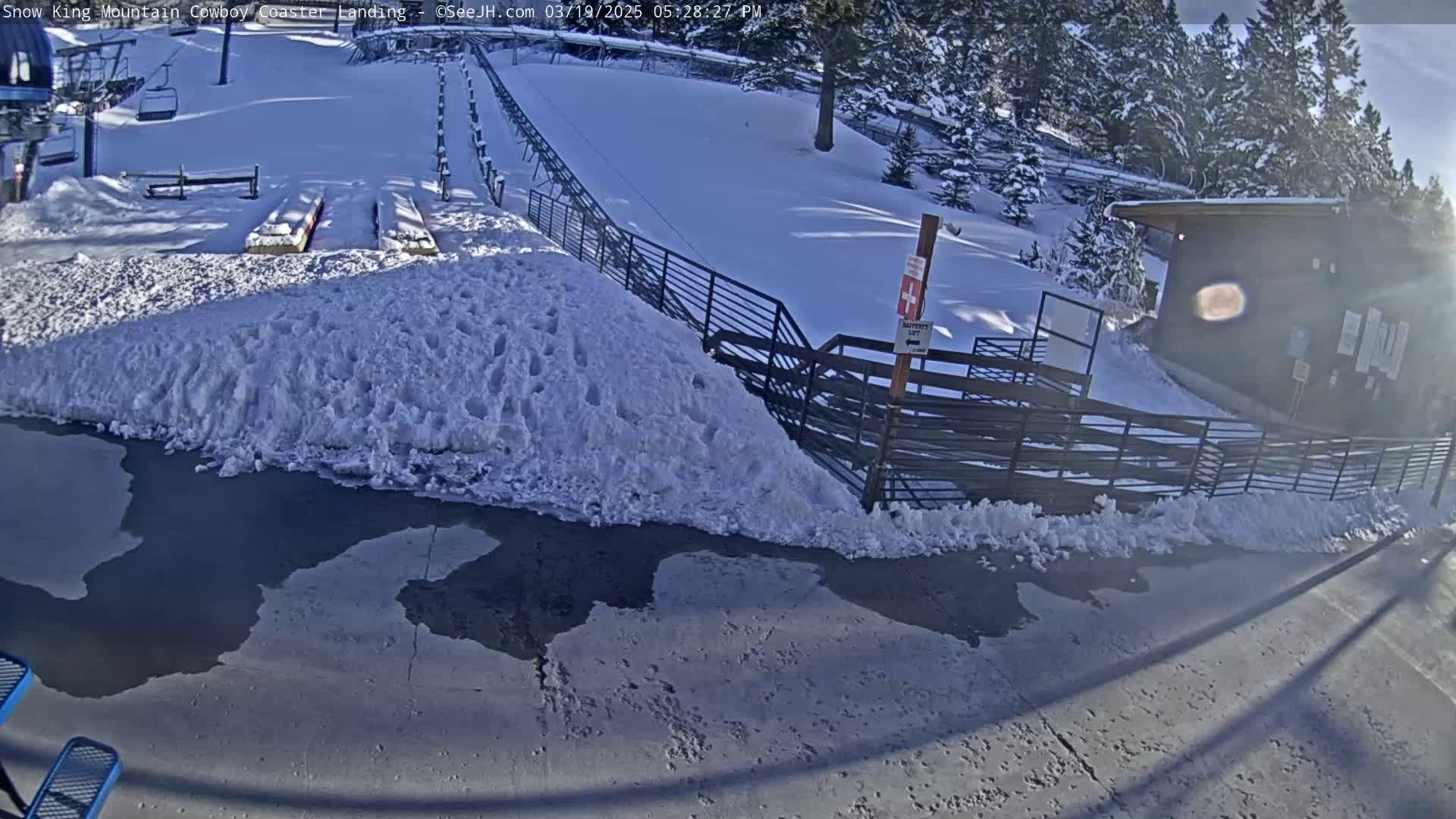 A snow-covered landing area for a mountain coaster features a metal fence, a small building, and patches of melting snow and ice on the pavement, all under a sunny sky.