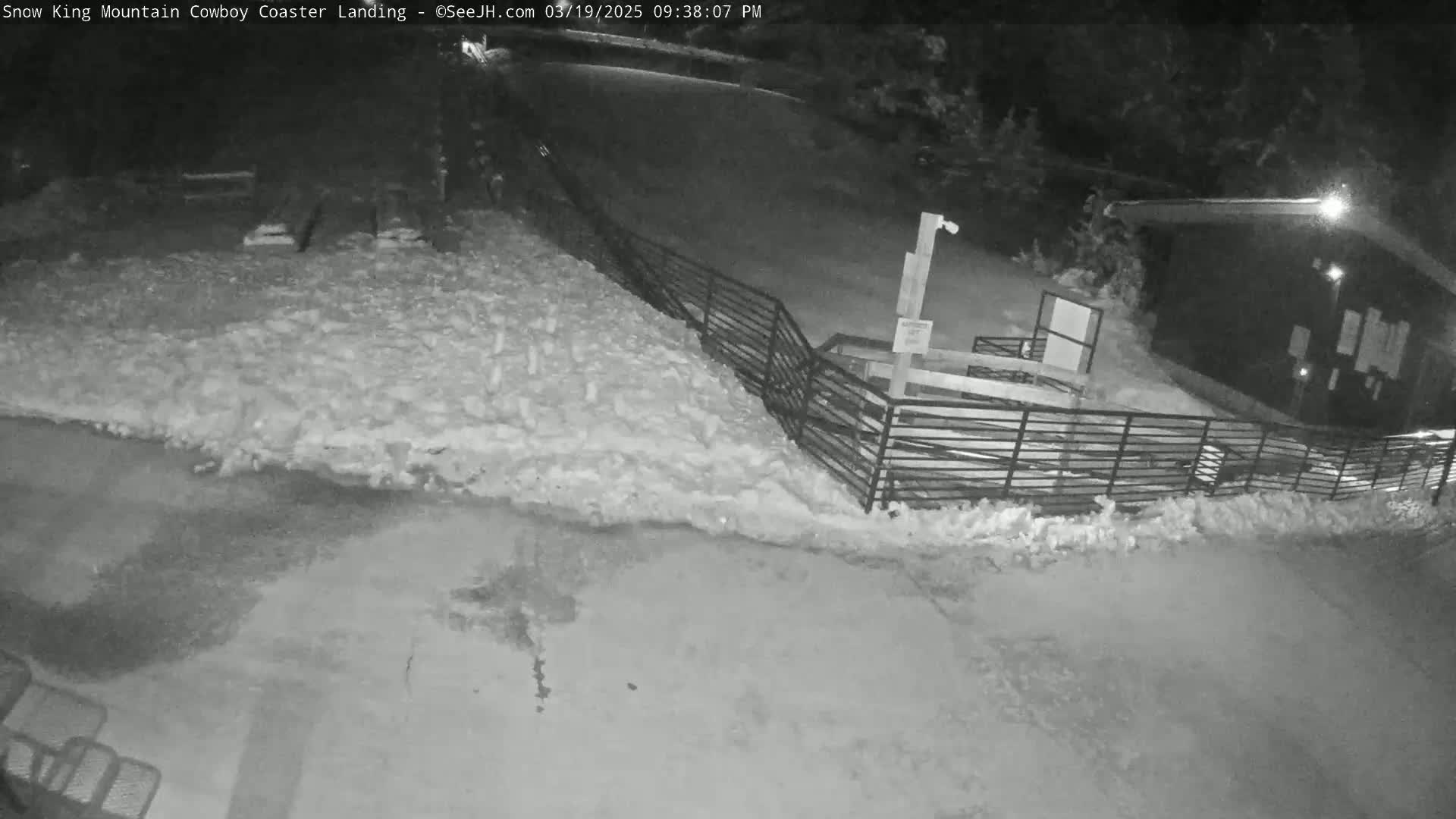 A nighttime, snow-covered scene shows a metal fence bordering a small building, with a snow-covered area extending to the left.