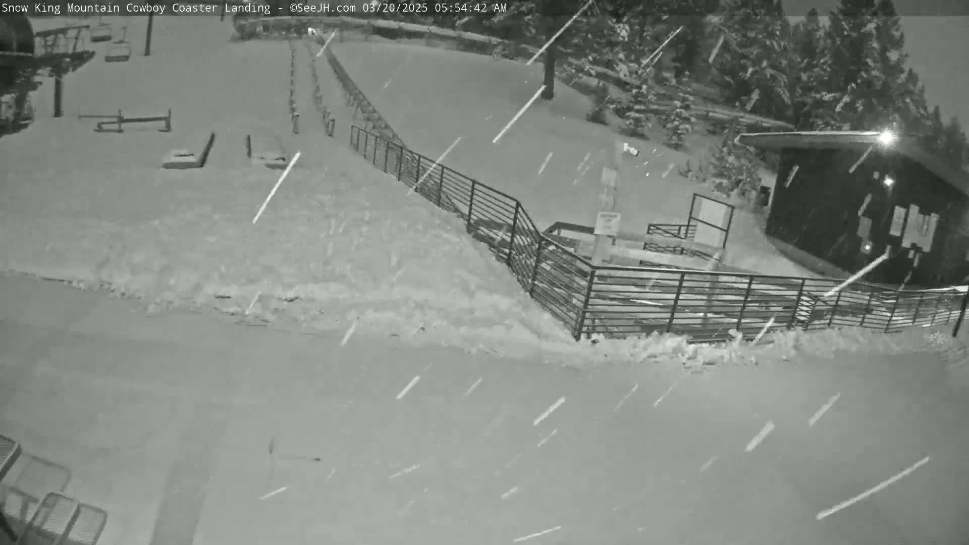 A snow-covered area with a small building and metal fences is seen during a nighttime snowfall.