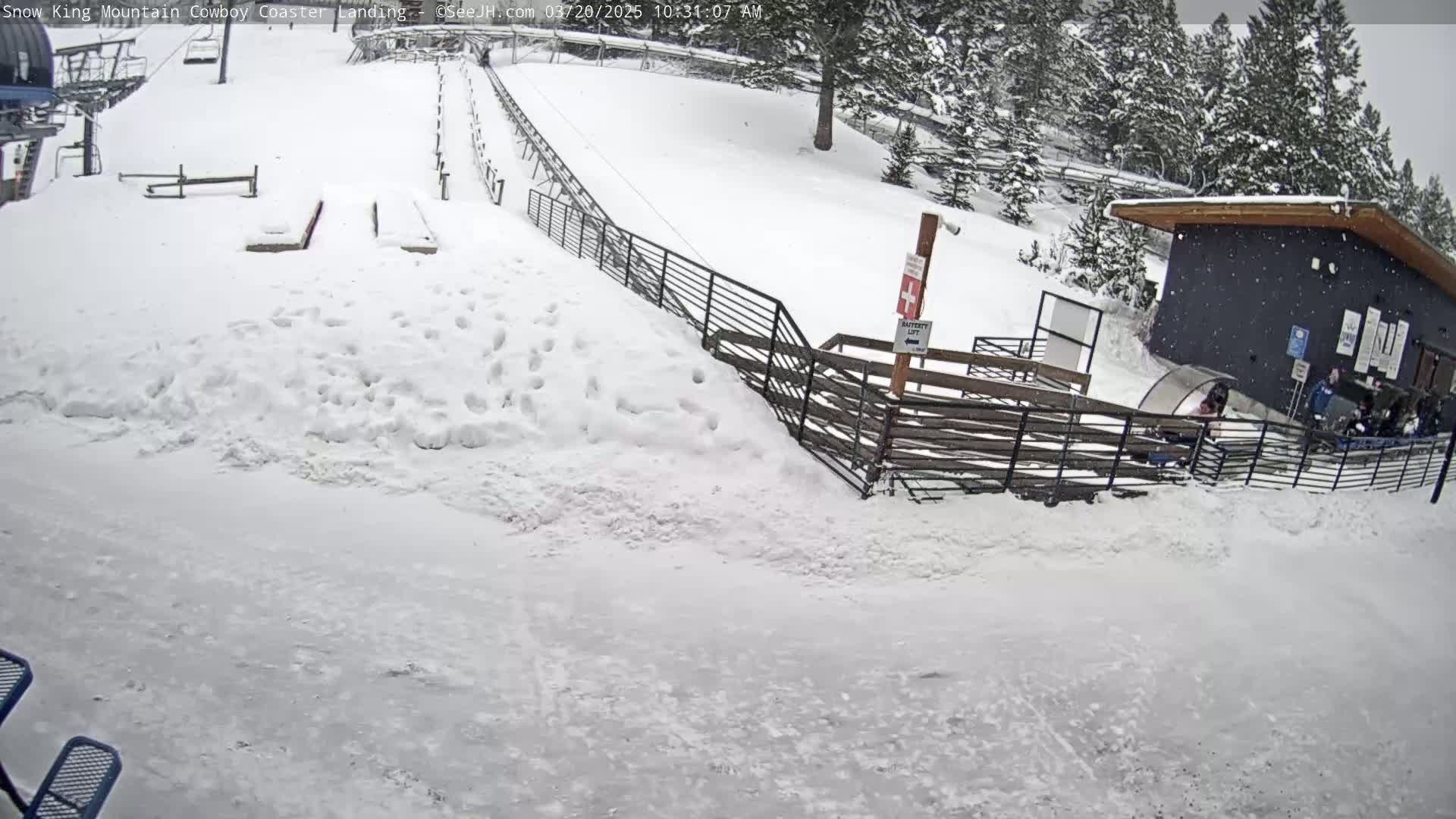 A snow-covered coaster track and landing area are seen during a snowfall, with a small building and people nearby.