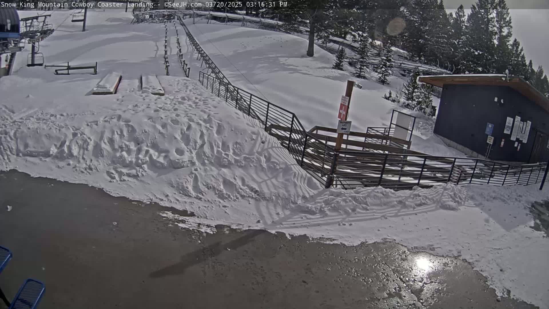 A snow-covered mountainside scene shows a coaster landing area with a small building and metal fences, under a partly sunny sky.