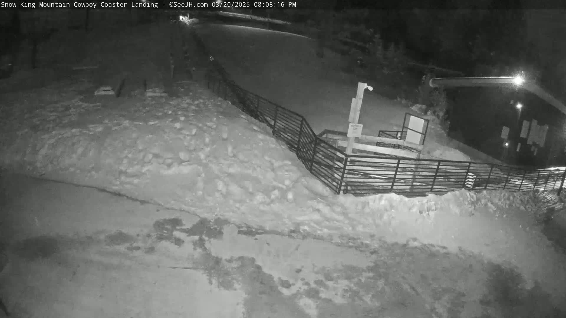 A snow-covered area at night, viewed from above, shows a metal fence leading to a dark building.