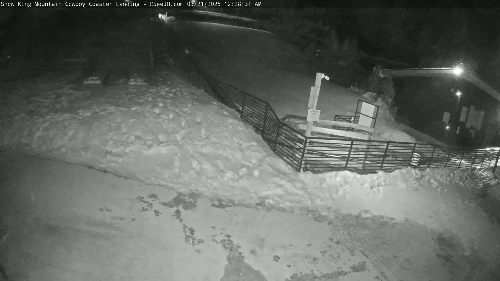 A snow-covered area at night, showing a dark building, a metal fence, and a sloped, snow-laden ground.