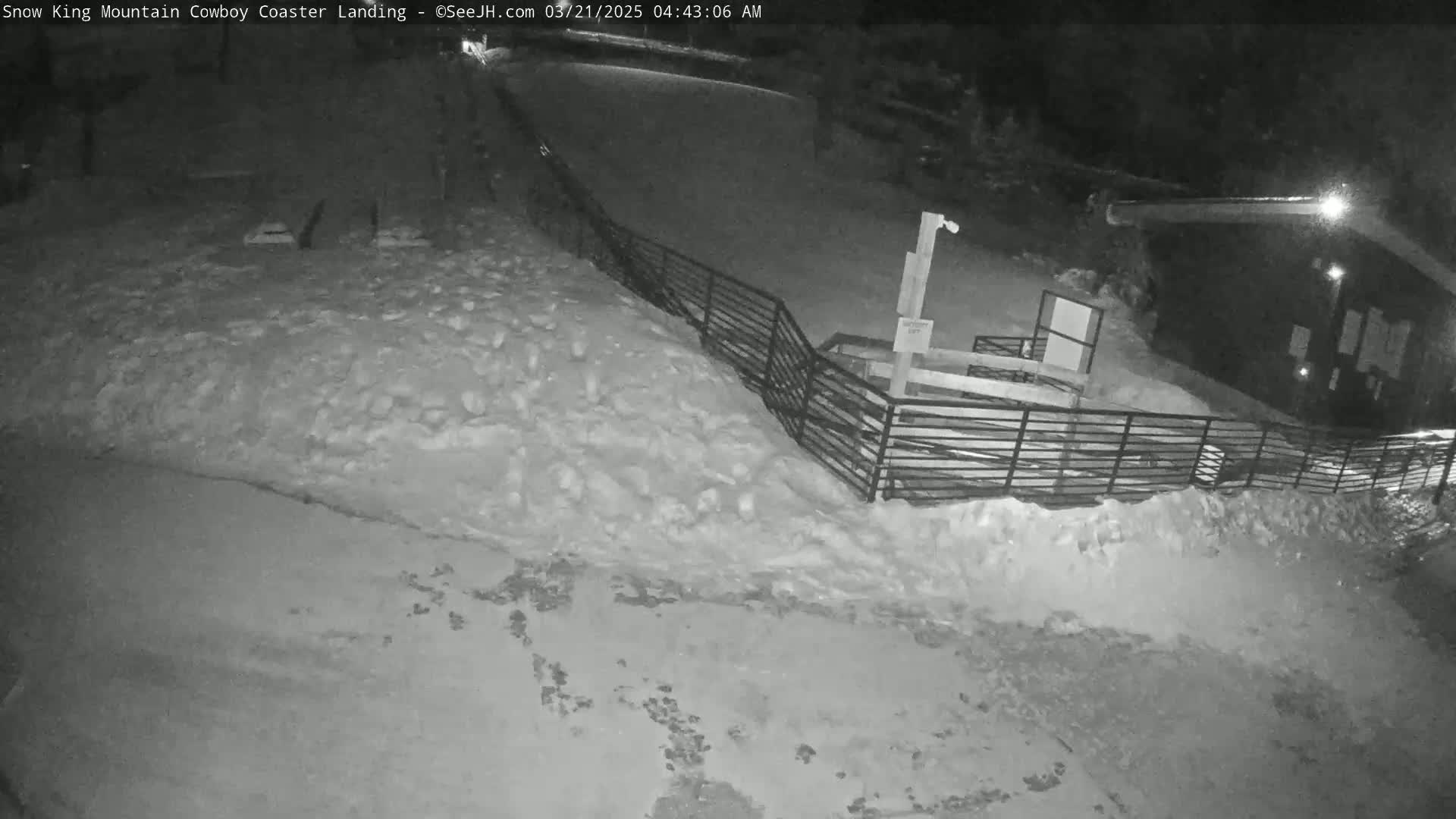 A snow-covered area at night, featuring a metal fence and a building in the background.