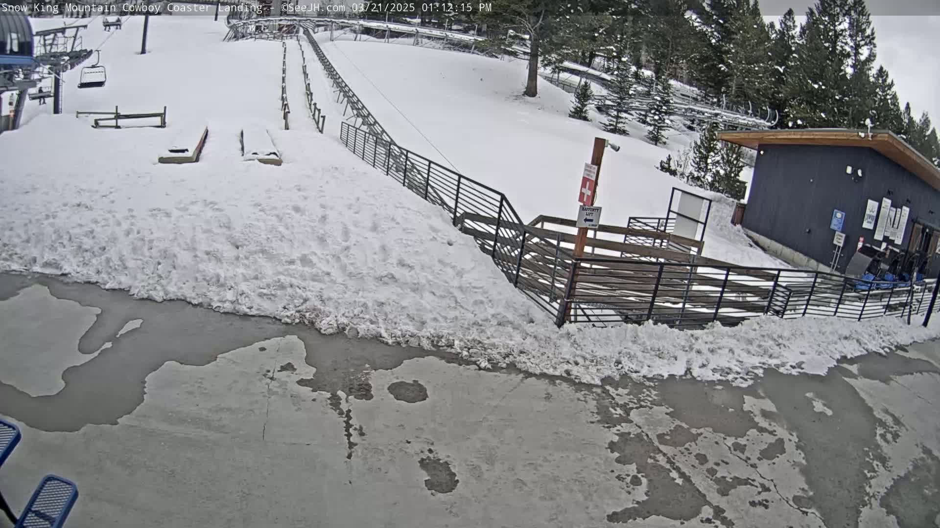 A snowy scene shows a coaster landing area with a small building, a metal fence, and melted patches of snow and ice on the ground.