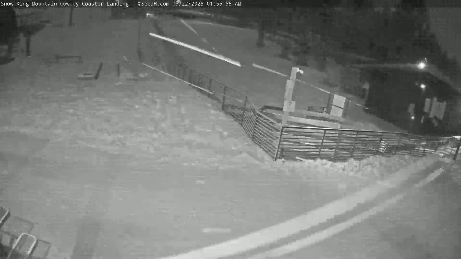 A nighttime, snowy scene shows a snow-covered area with a metal fence, a building in the background, and faint vehicle headlights visible on a road.