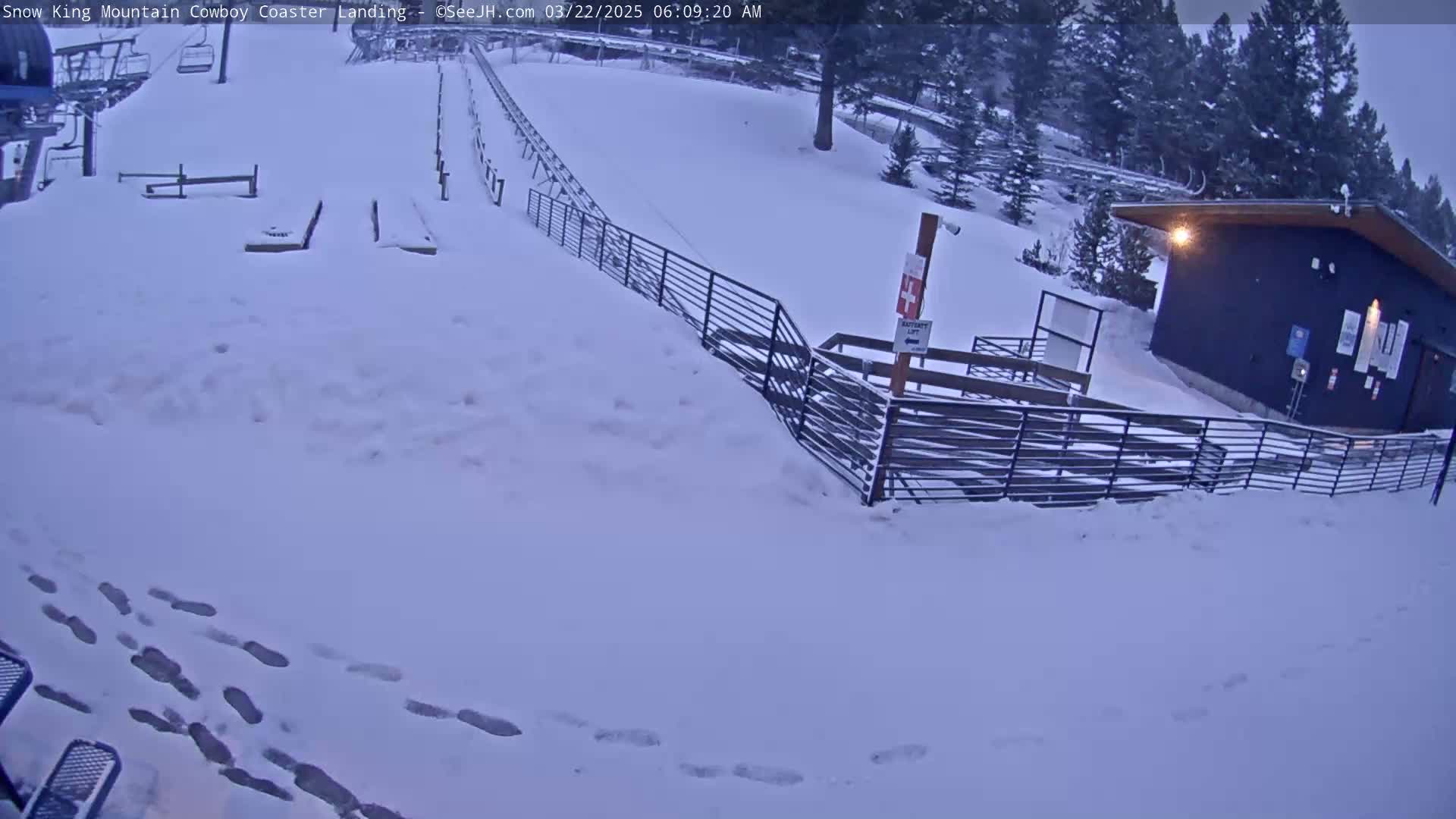 A snow-covered mountainside shows a coaster landing area with a small building and footprints in the snow.