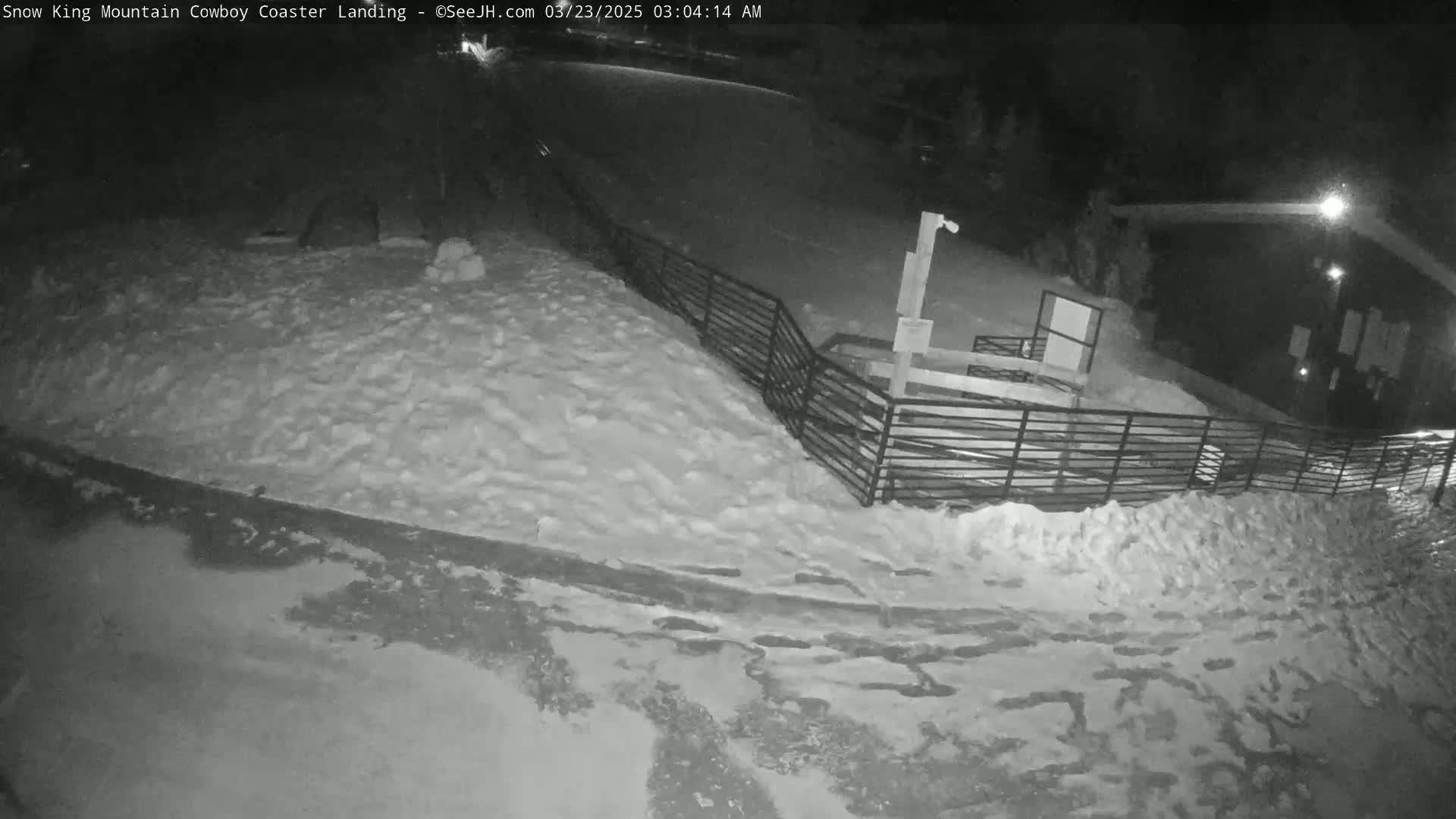 A snow-covered area at night, with a metal fence and a building visible in the background.