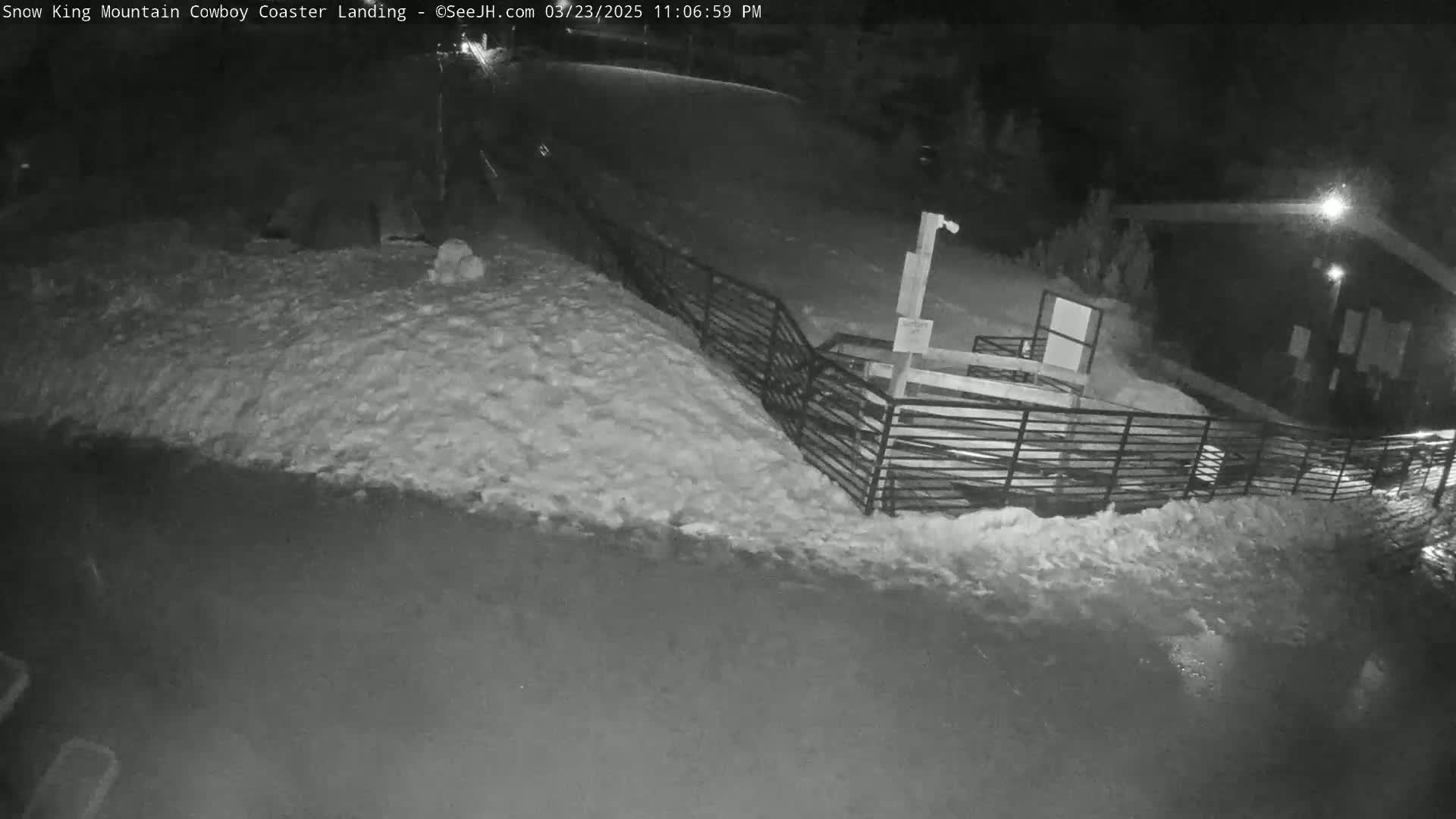 A snow-covered area at night is seen with a metal fence and a small building in the background.