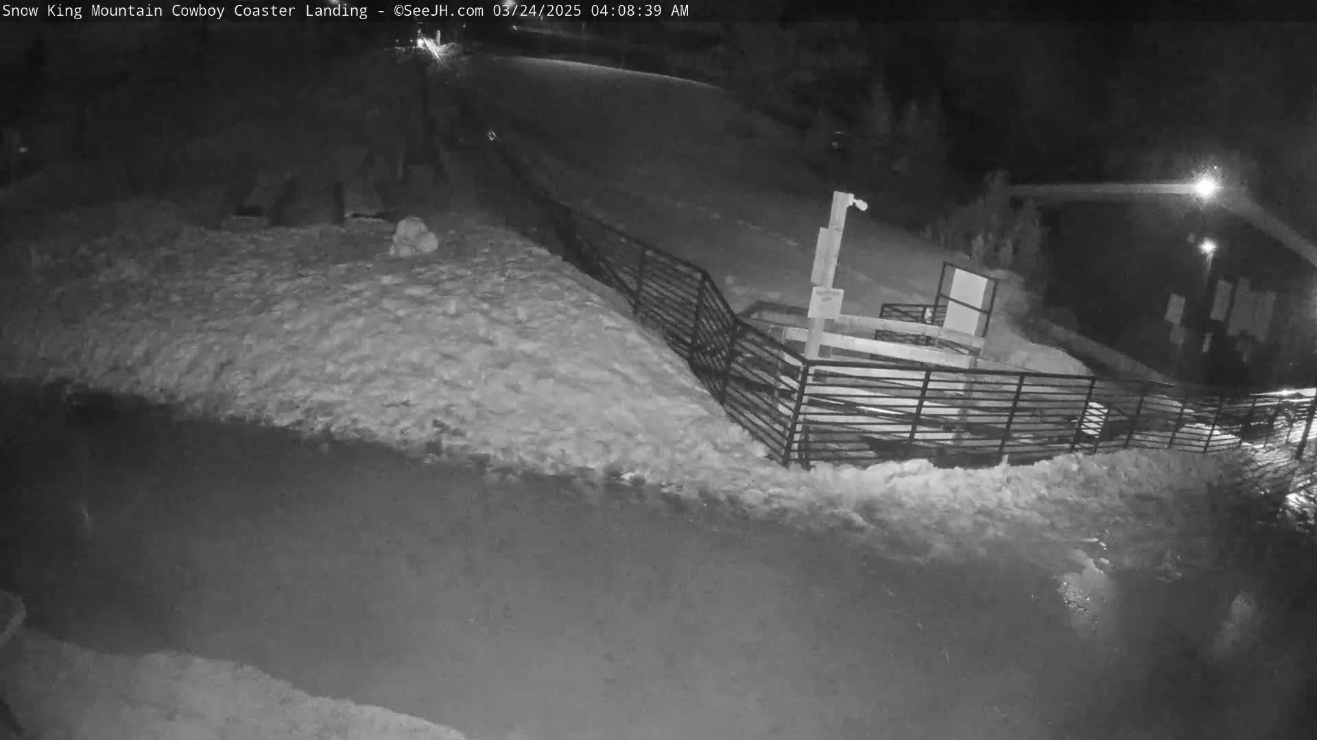 A nighttime, snow-covered scene shows a metal fence next to a dark building, with a snow-covered path leading away.