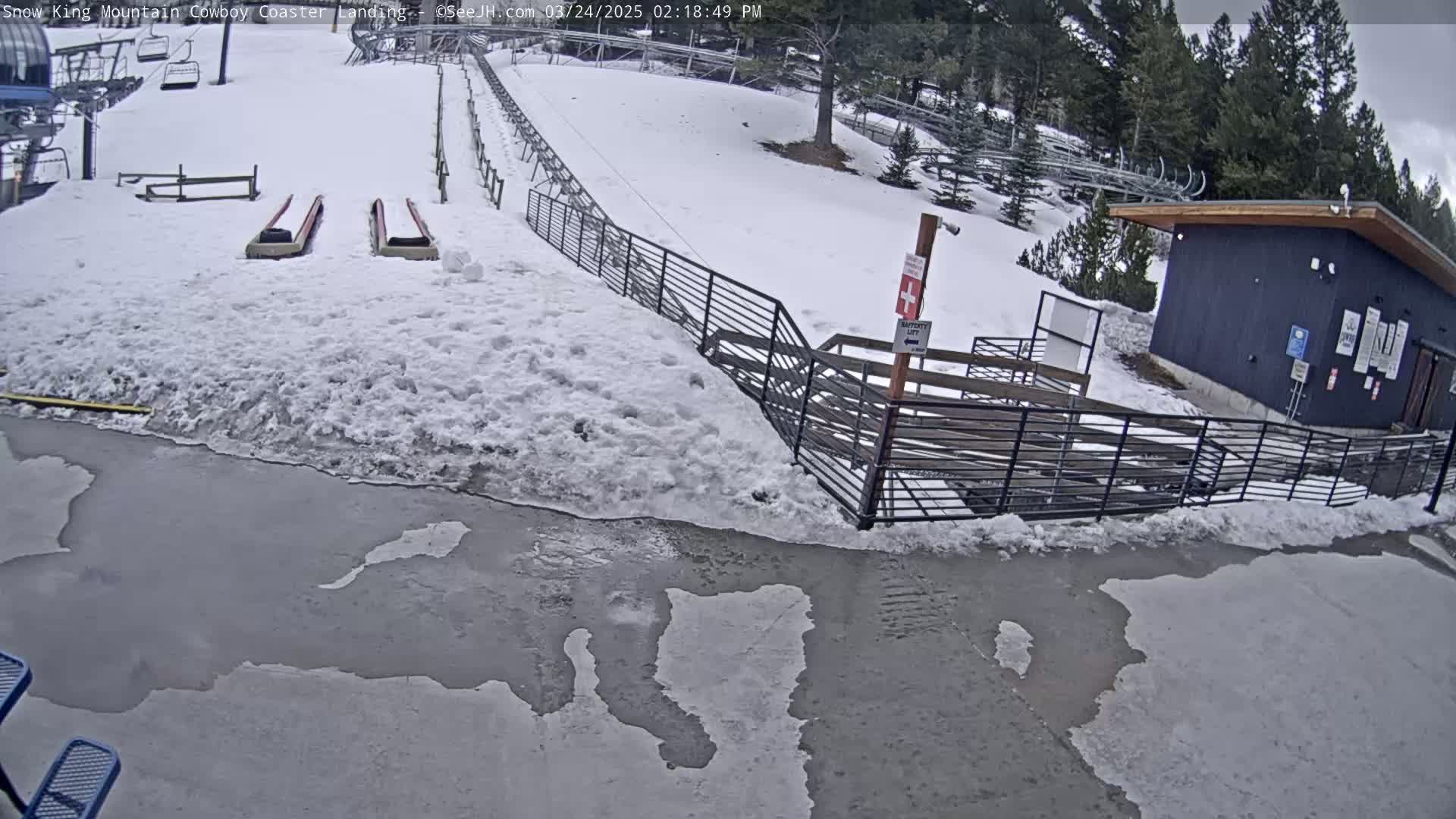 A snowy mountainside scene shows a snow-covered landing area for a mountain coaster, a small building, and a partially melted ice patch on the ground.