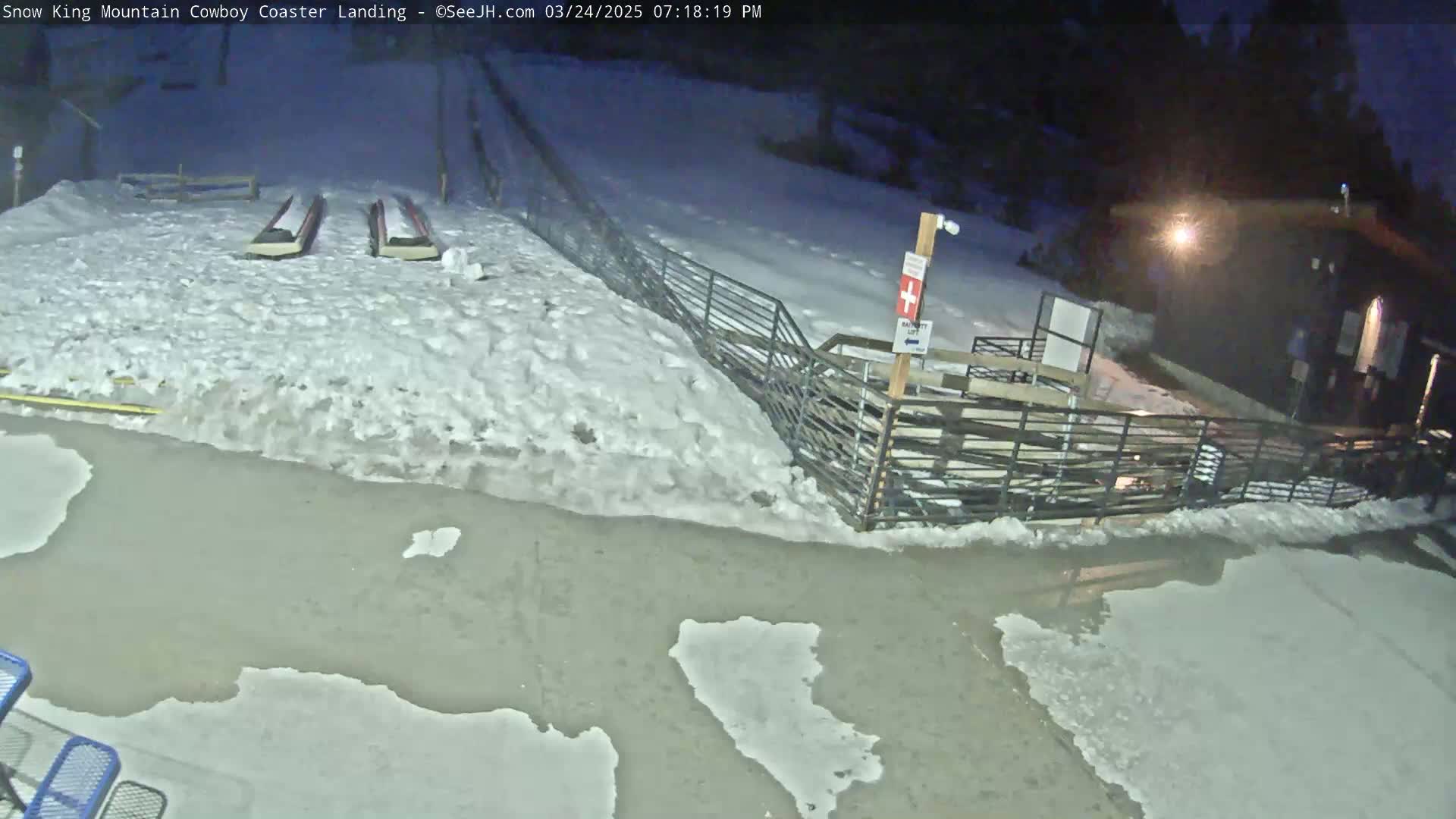 A nighttime scene shows a snow-covered area with patches of melted snow and ice, two long sleds, and a metal fence near a building.