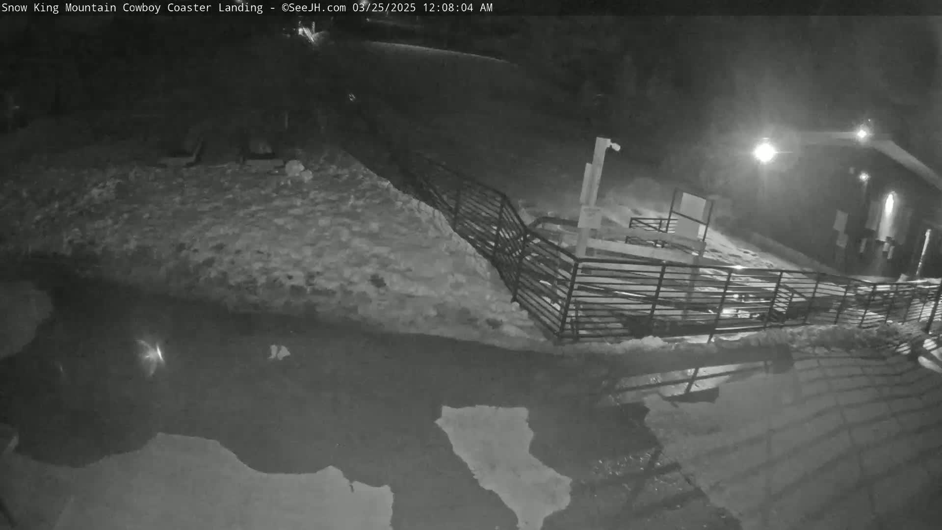 A nighttime, snow-covered area with a metal fence and puddles of water is illuminated by artificial light.