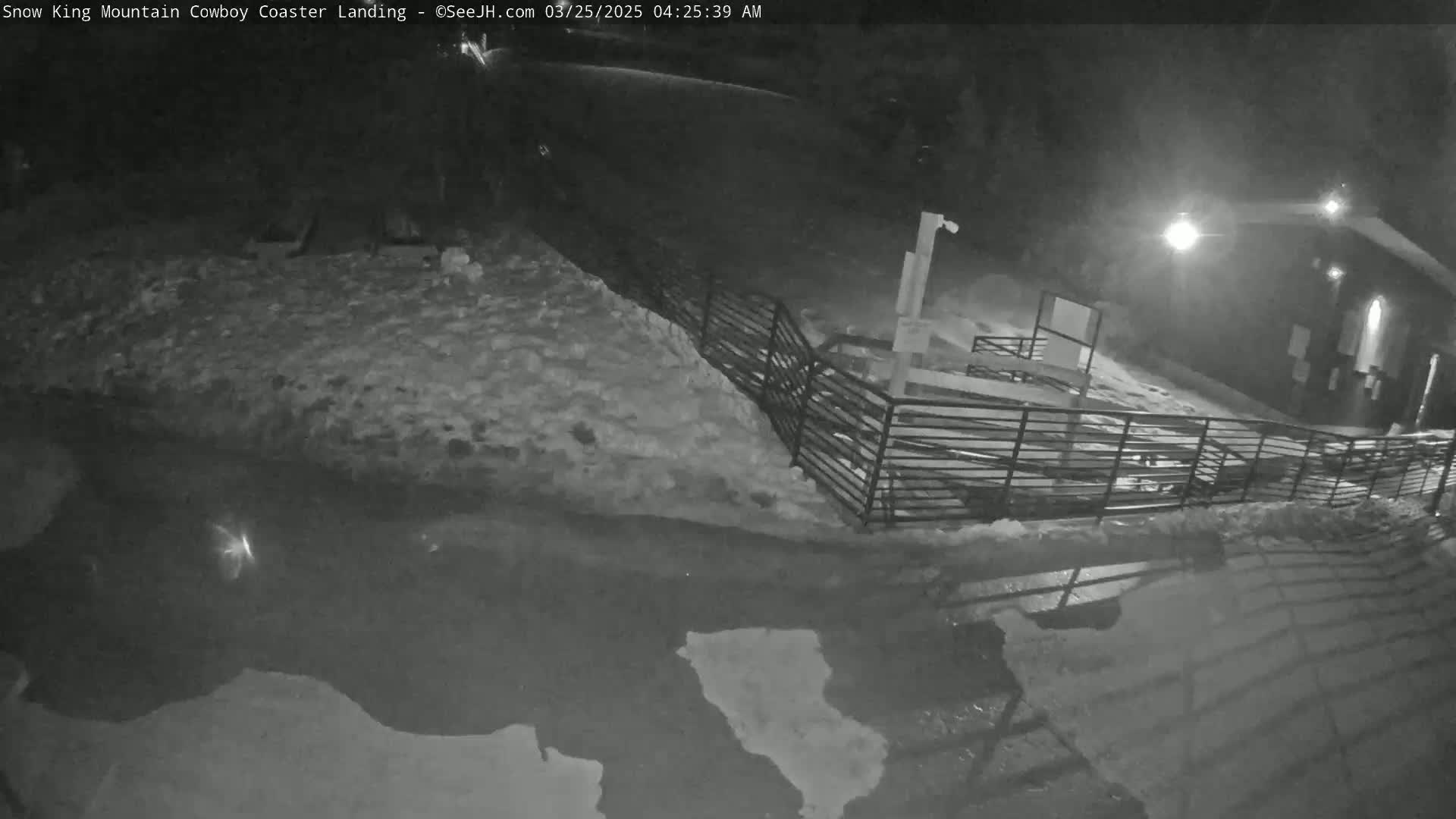 A nighttime, snow-covered scene shows a metal fence surrounding a paved area with puddles, illuminated by artificial lights.