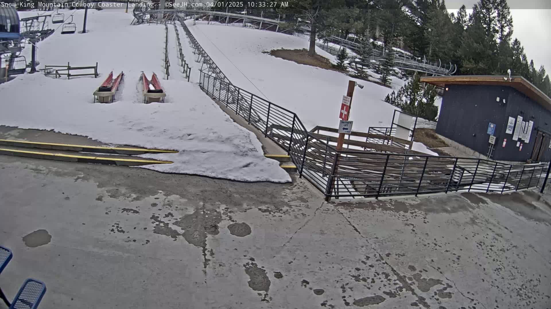 A snowy outdoor area shows a landing for a mountain coaster with two empty carts, a nearby building, and metal railings leading to steps.
