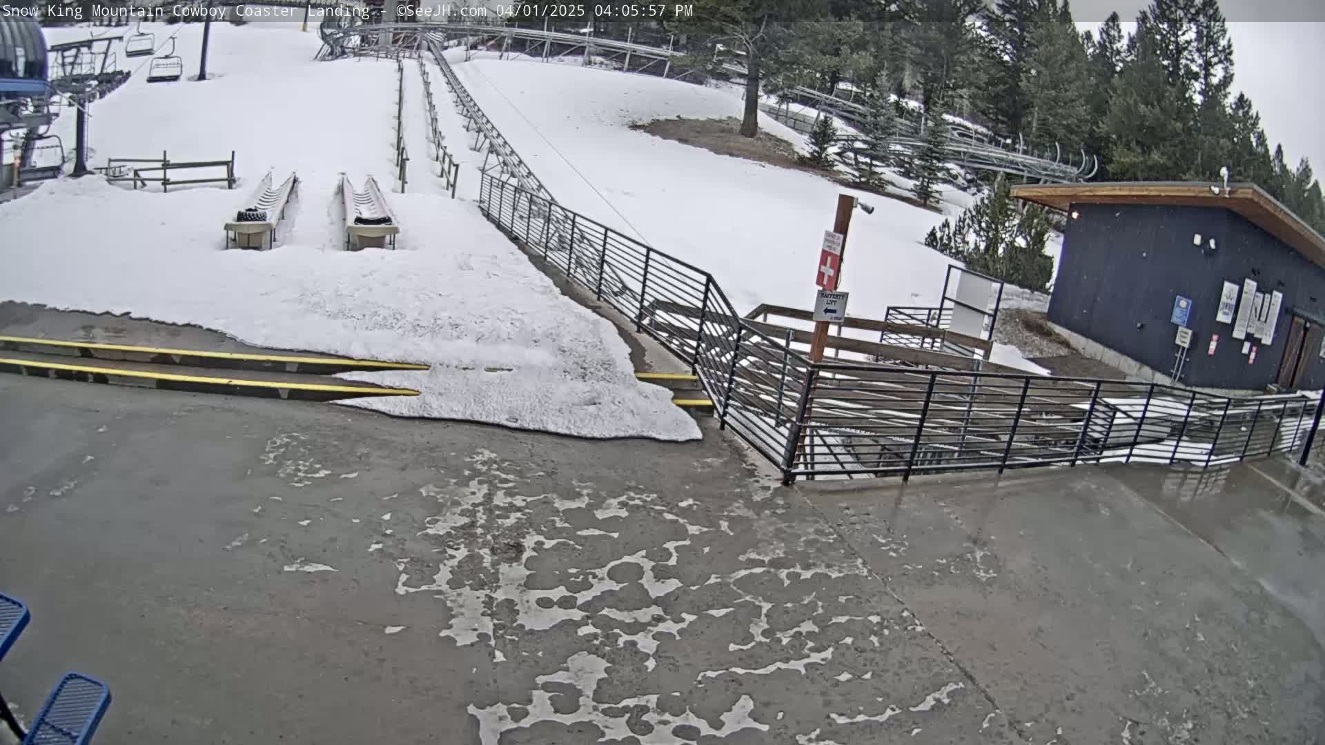 A snowy area with a coaster landing, metal railings, and a dark-colored building, with melting snow and wet pavement.