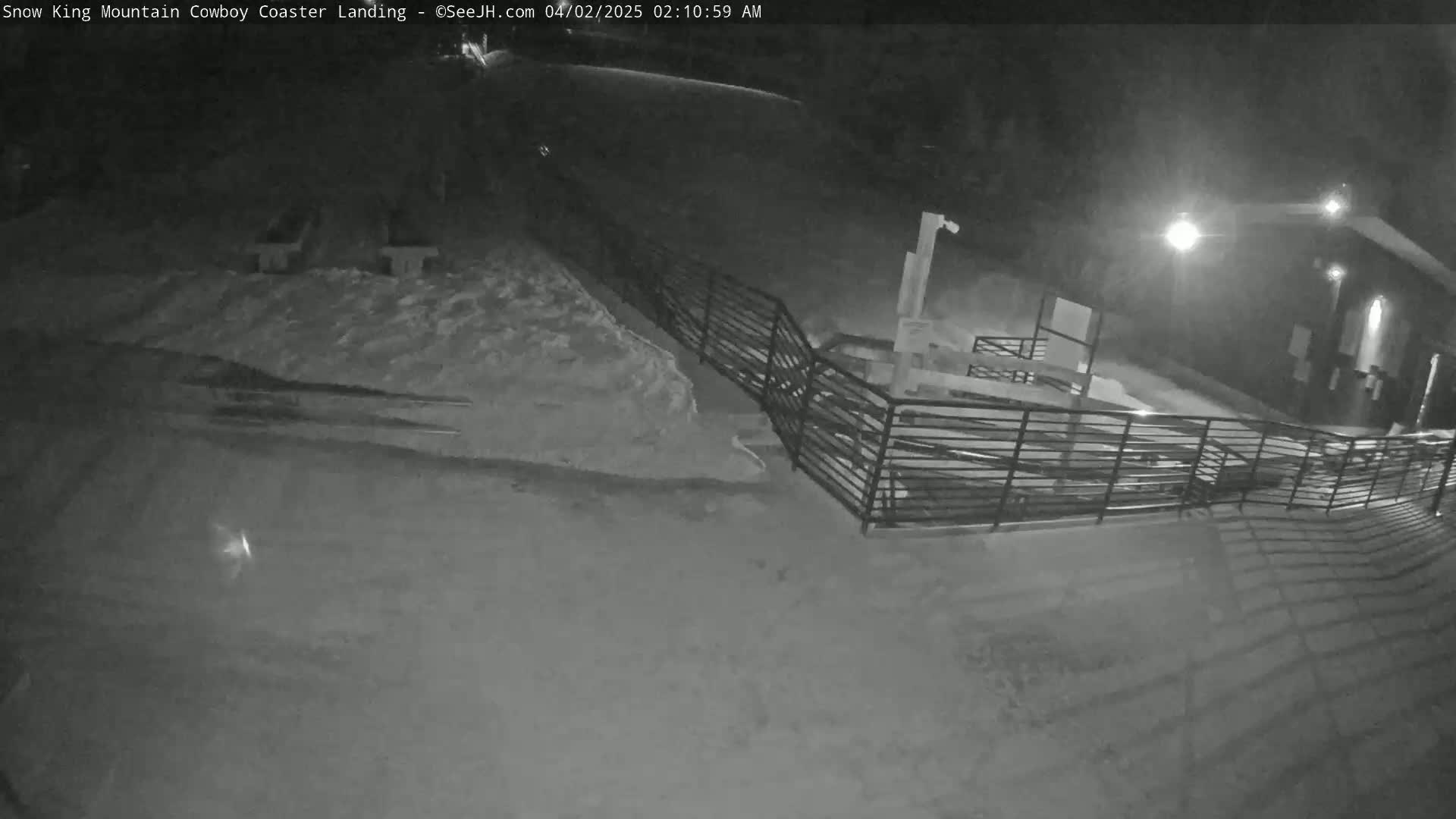 A nighttime, snow-covered area with a metal railing and a building in the background under artificial light.