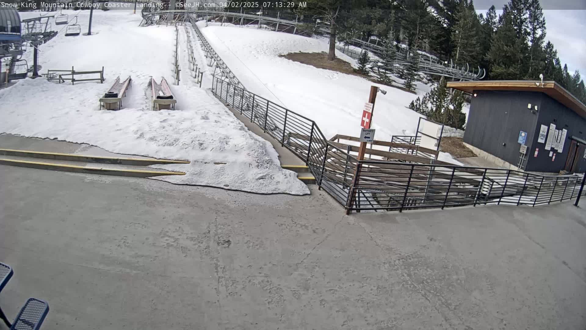 A snow-covered mountainside scene shows a coaster landing area with snow-covered ramps, metal railings, a small building, and a partly cloudy sky.