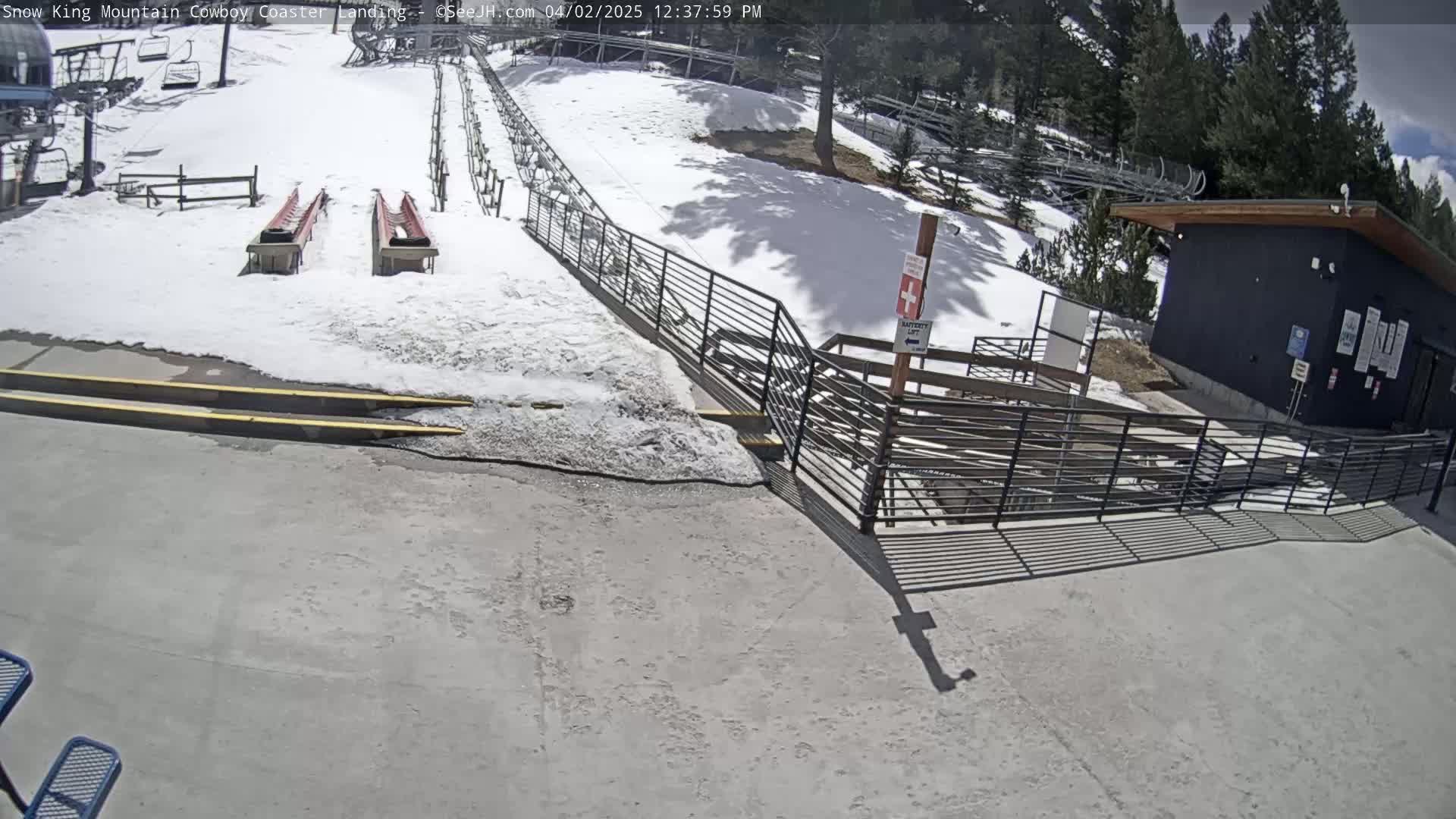 A snow-covered area with two empty red sleds, a metal railing, and a dark building under a partly sunny sky.