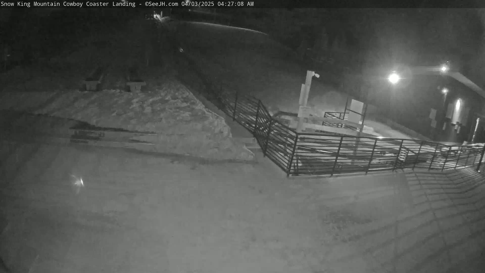 A snow-covered nighttime scene shows a metal railing near a building, with a path leading away from the camera.