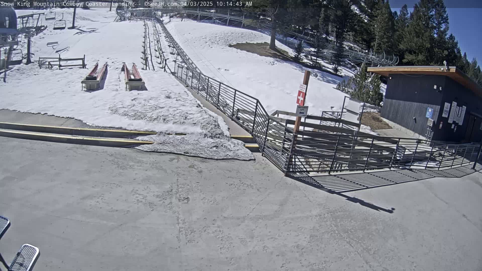 A snow-covered landing area for a coaster, with a nearby building and metal railings, under sunny skies.