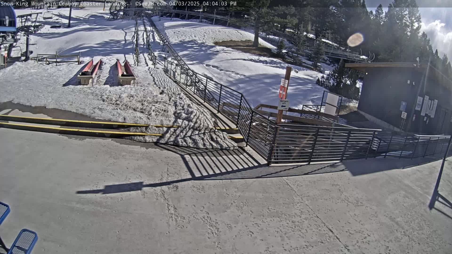 A snowy outdoor scene shows a coaster landing area with two red sleds, metal railings, and a small building under a partly sunny sky.