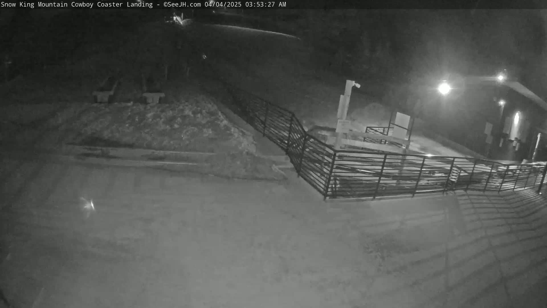 A nighttime, snow-covered area shows a metal railing leading to a building under artificial light.