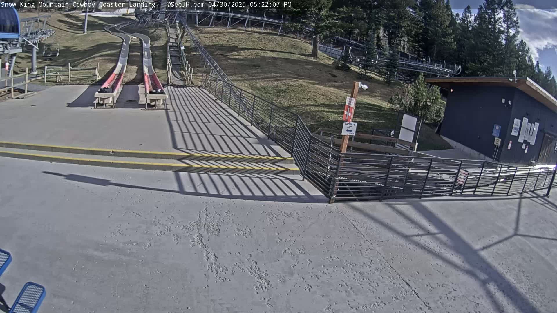 Two snow-coaster tracks end at a paved landing area with stairs and a fence, under a mostly sunny sky.