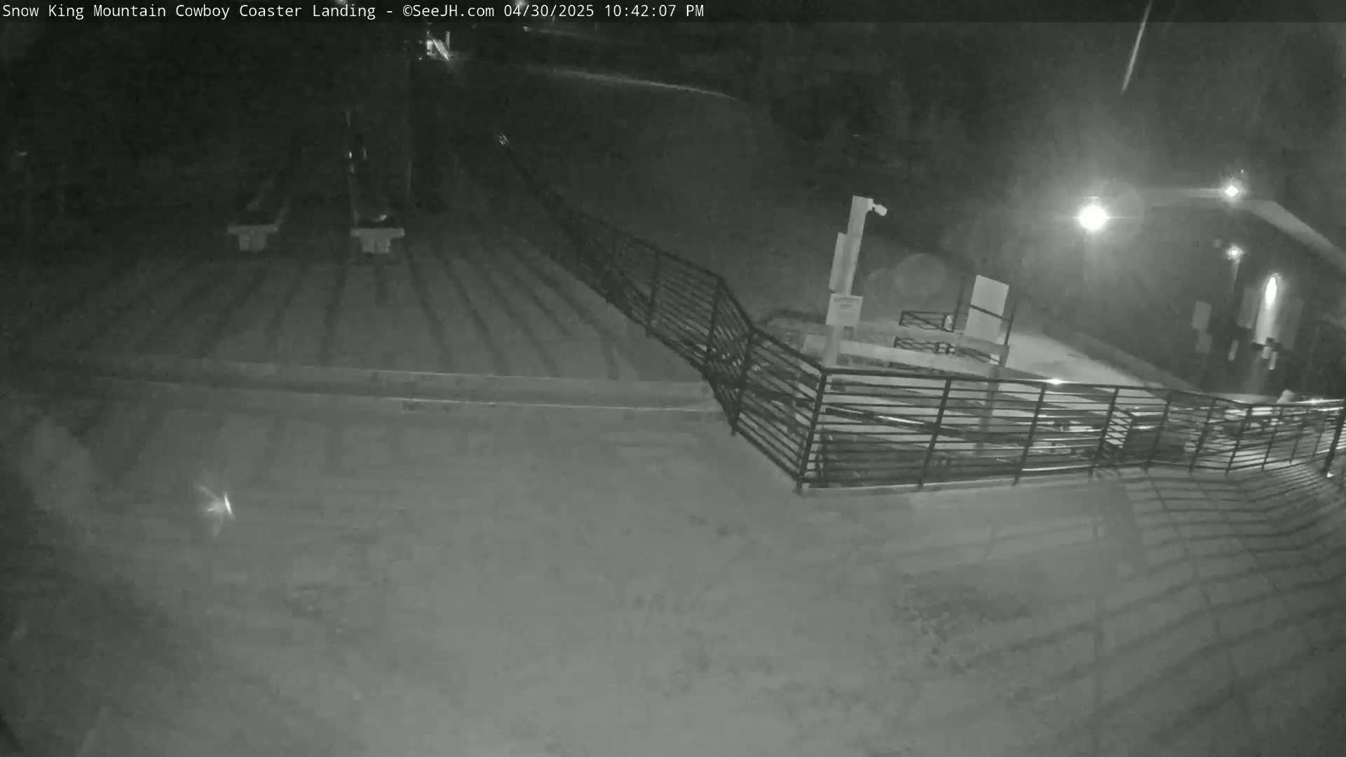 A nighttime, snow-covered area with a metal railing and some structures visible under artificial light.